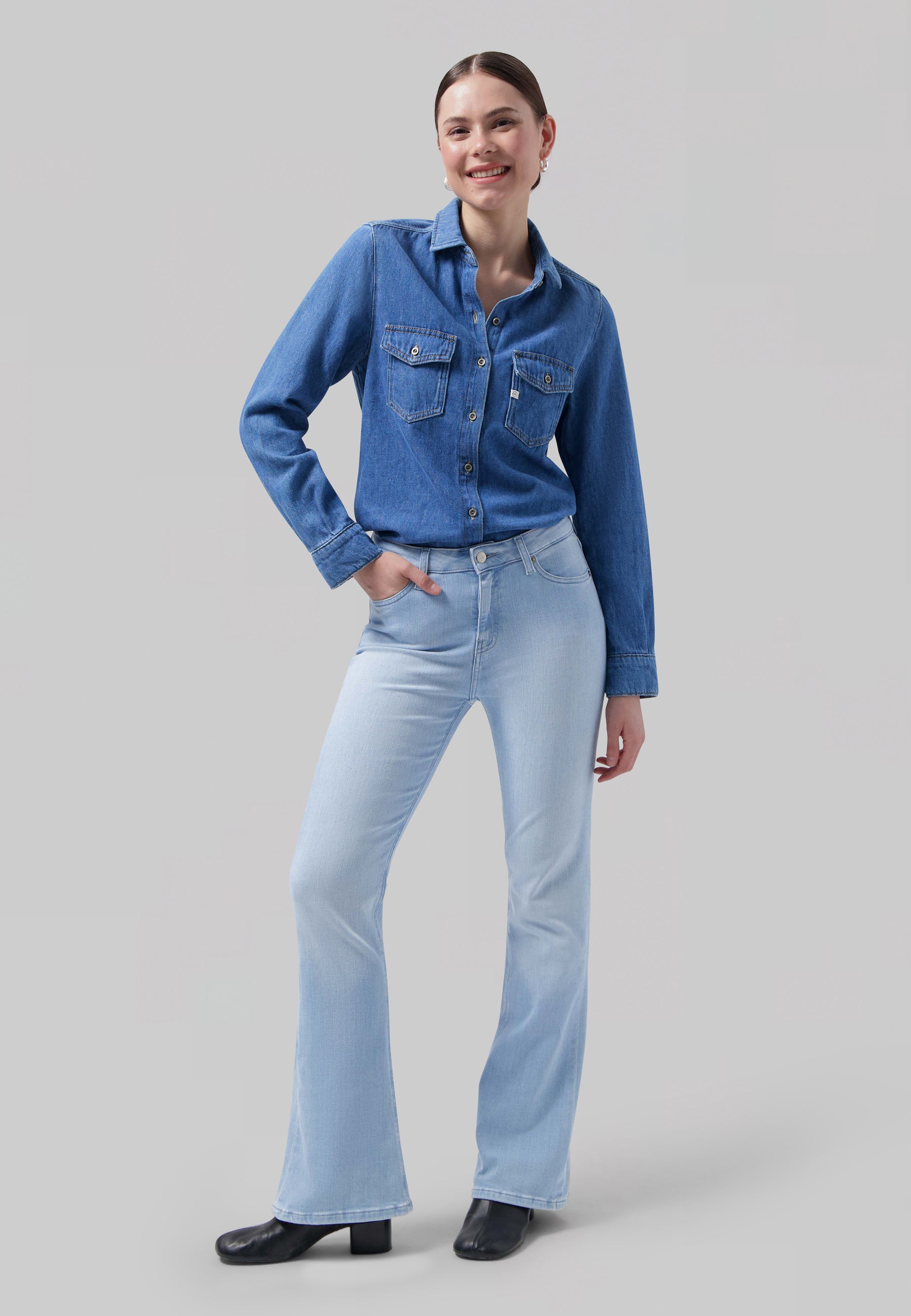 A woman wearing a classic blue denim shirt with front pockets paired with light blue flared jeans and black ankle boots.