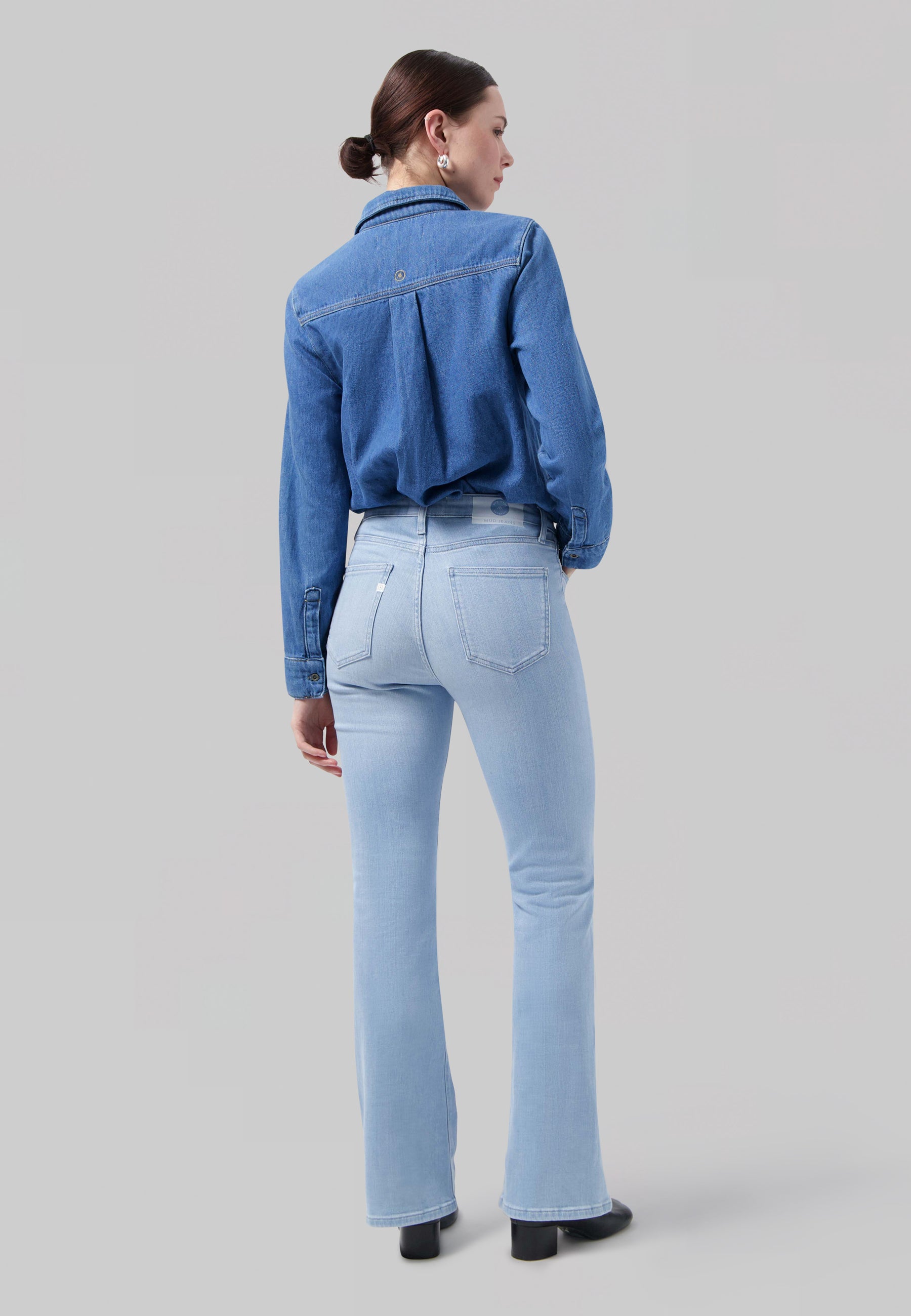 Woman wearing light blue flared jeans and a long-sleeve denim shirt, viewed from the back, styled with earrings.