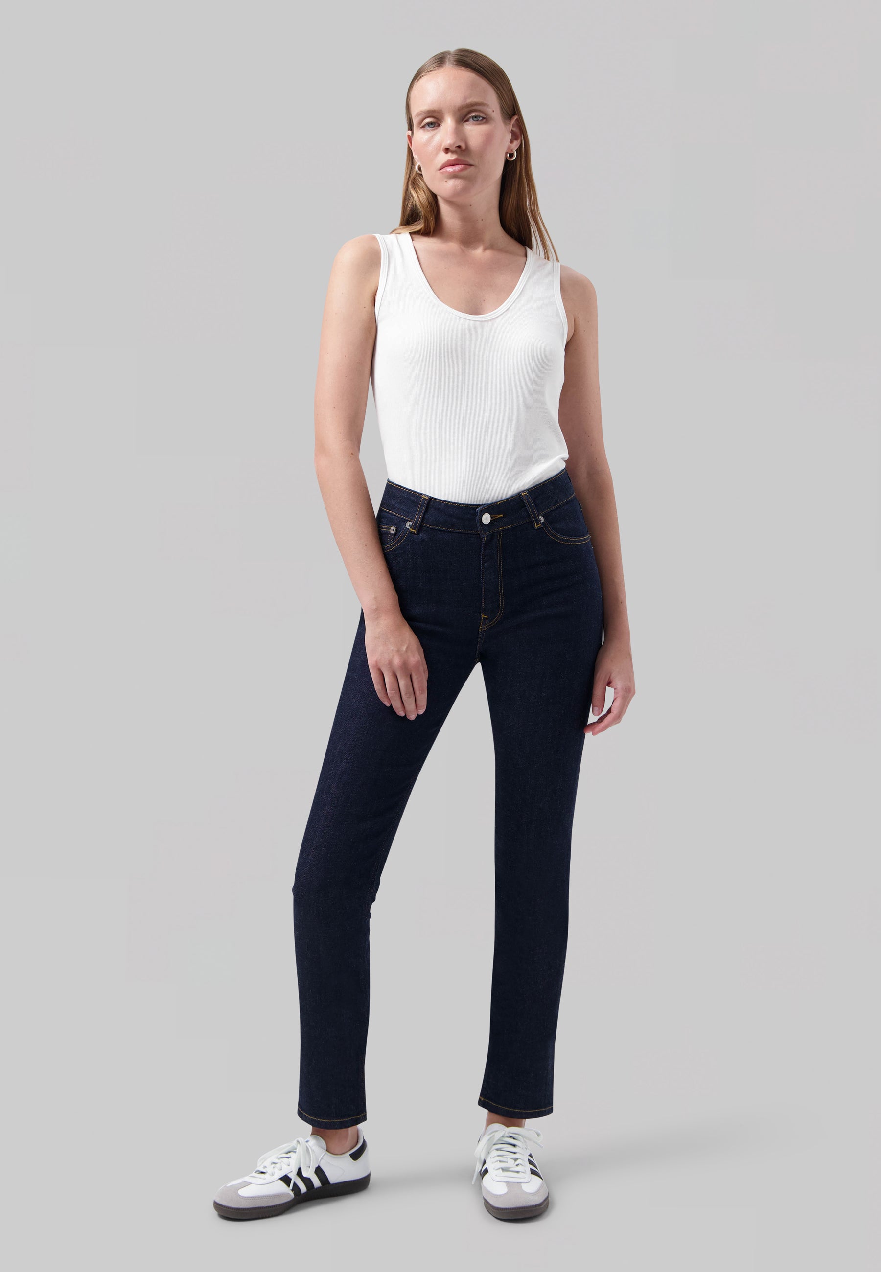 Woman modeling fitted dark blue jeans paired with a white sleeveless top, standing against a plain gray background.