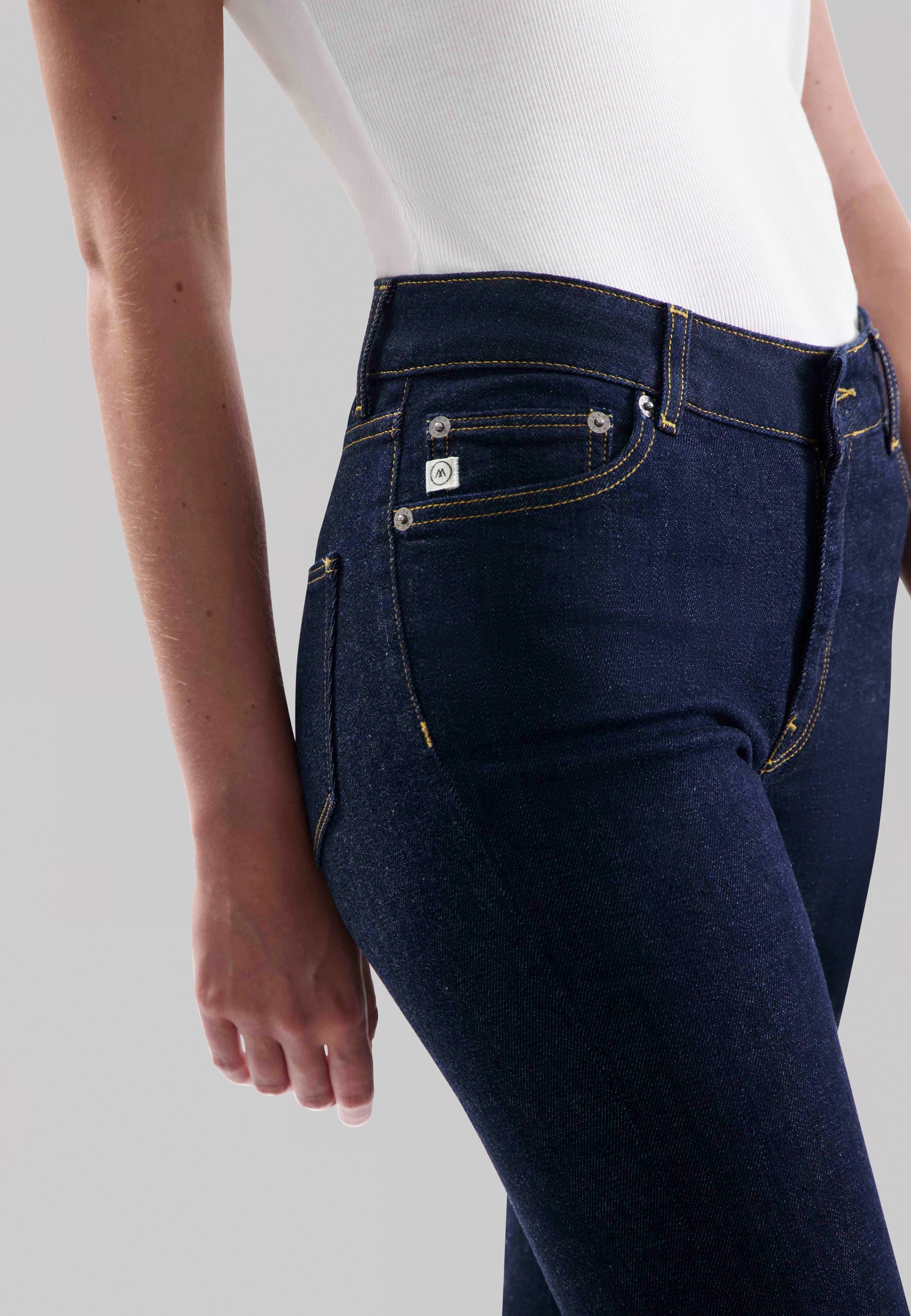 Close-up of dark indigo jeans with contrast stitching worn by model in a fitted white ribbed tank top, highlighting pocket details.