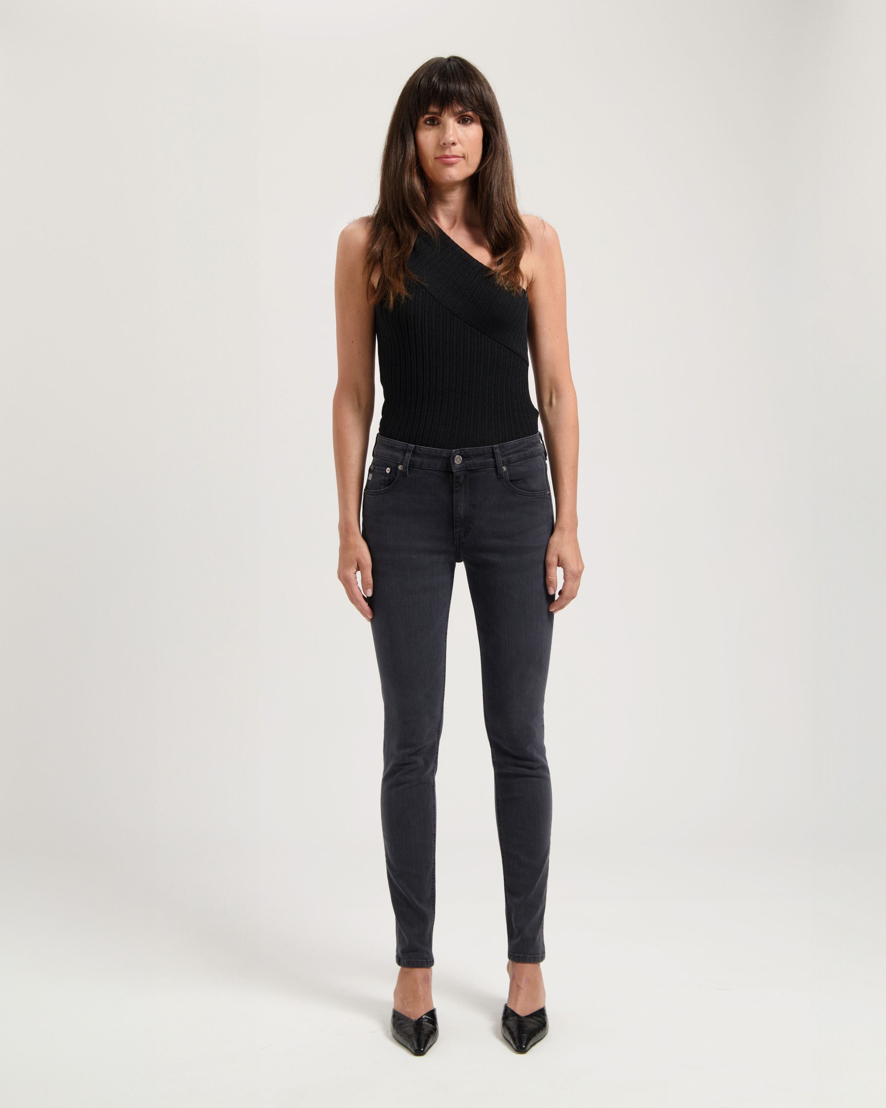 Woman wearing a stylish one-shoulder black ribbed top paired with dark gray skinny jeans and black pointed heels.
