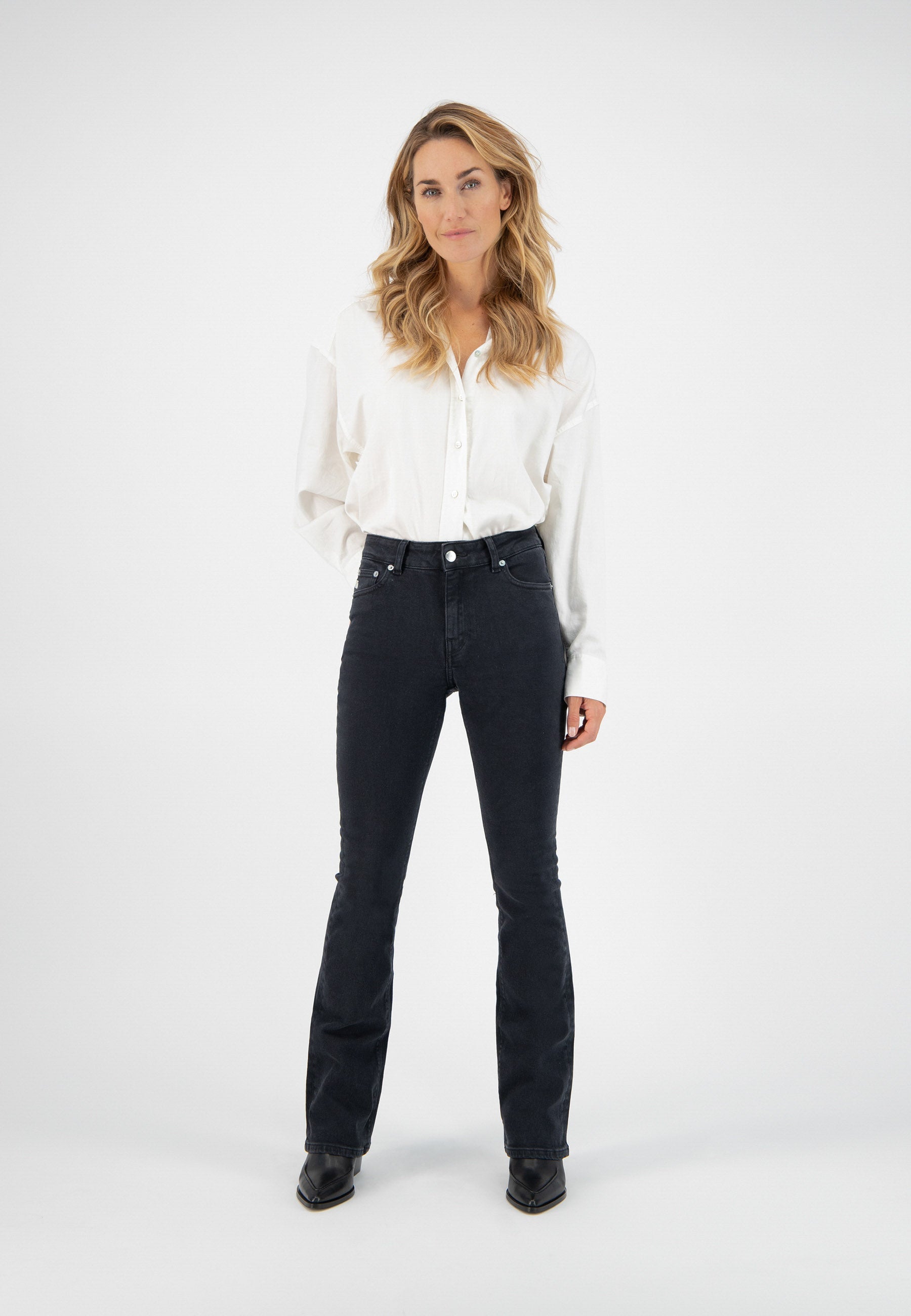 A woman wearing a white button-up blouse and black flared jeans paired with black pointed-toe boots against a plain background.