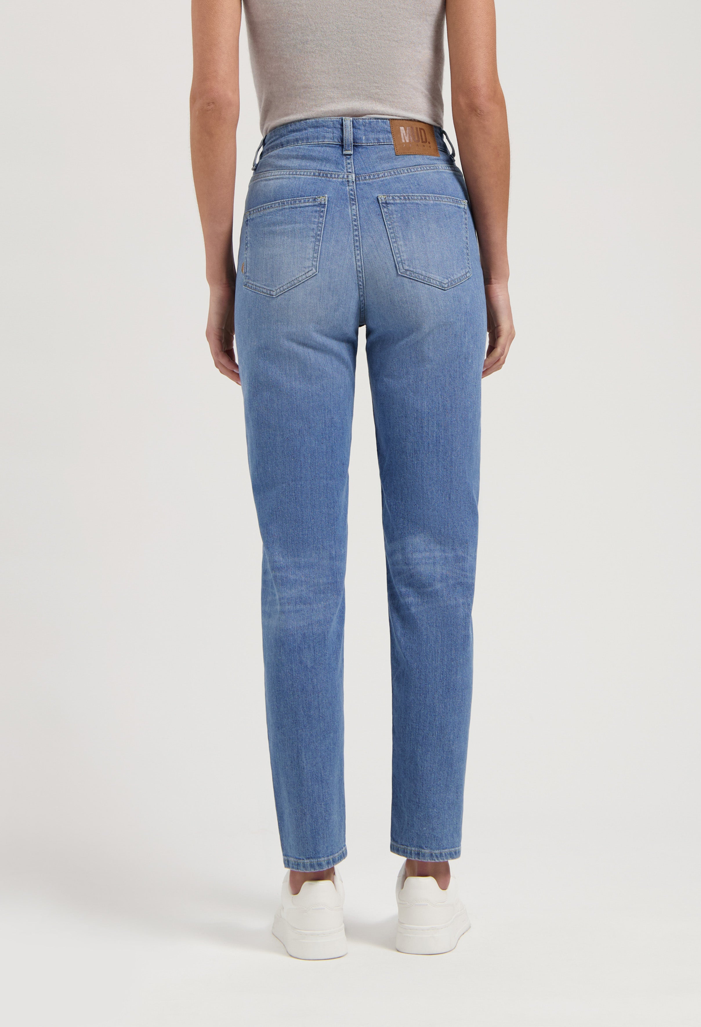 Mid-blue high-waisted jeans by MUD Jeans, featuring a tag on the waistband, paired with a gray sleeveless top and white sneakers.