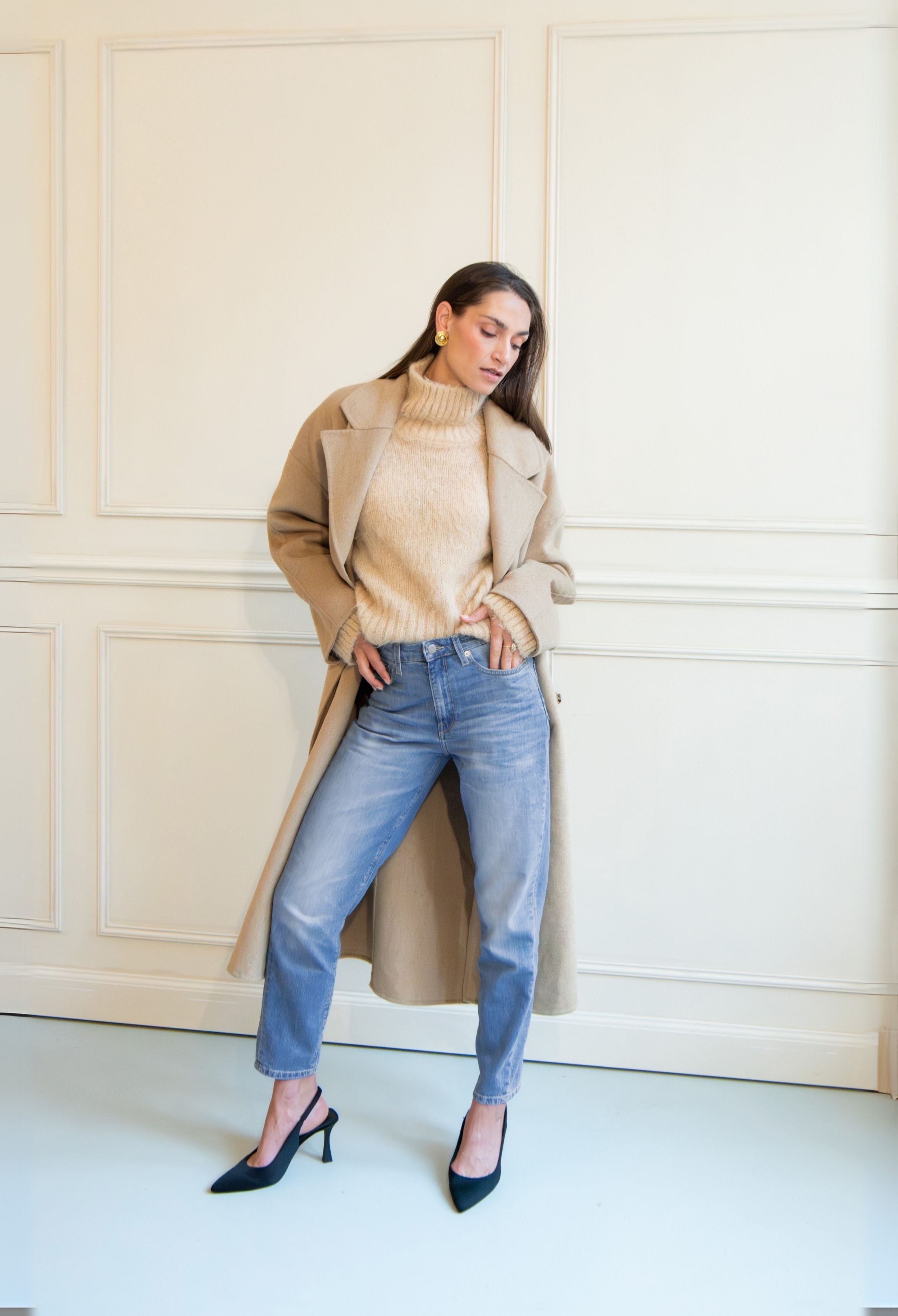 Stylish outfit featuring a light brown wool coat over a beige turtleneck sweater with light blue denim jeans and black heels.