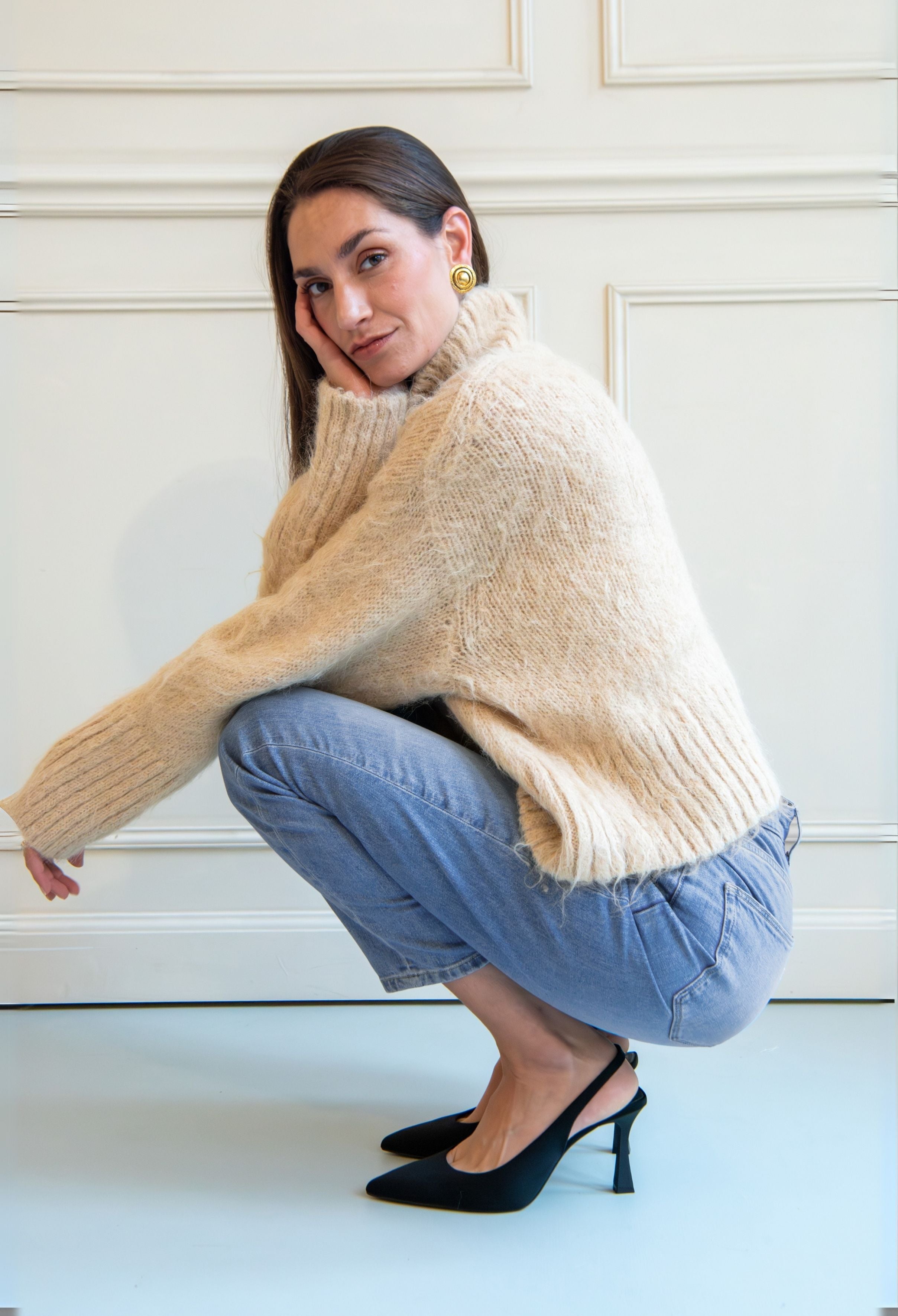 Woman wearing a cozy beige knitted sweater with light blue jeans and black pointed heels against a white paneled wall.