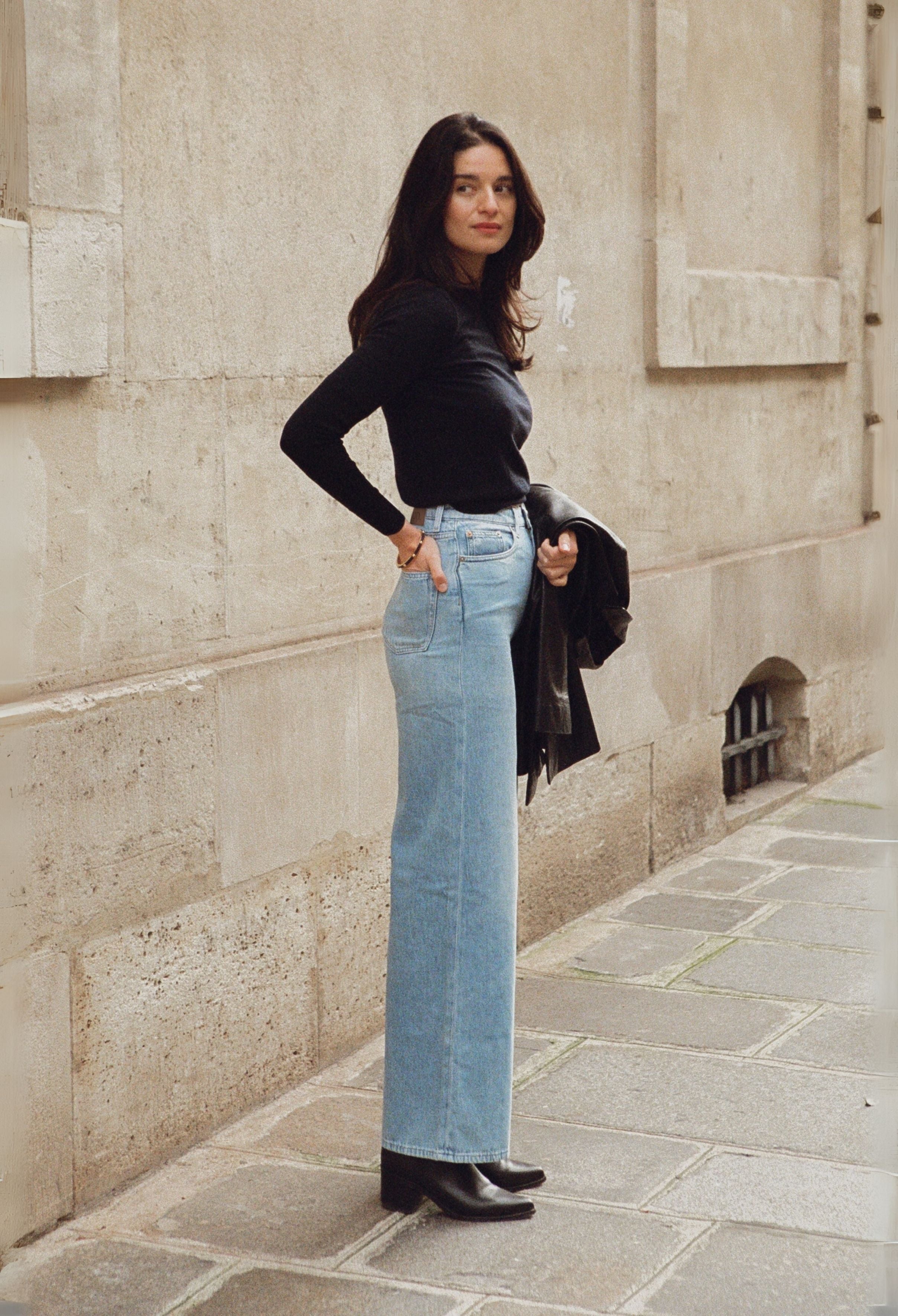Stylish woman in light blue high-waisted jeans and black top leans against a textured beige wall on a cobblestone street.