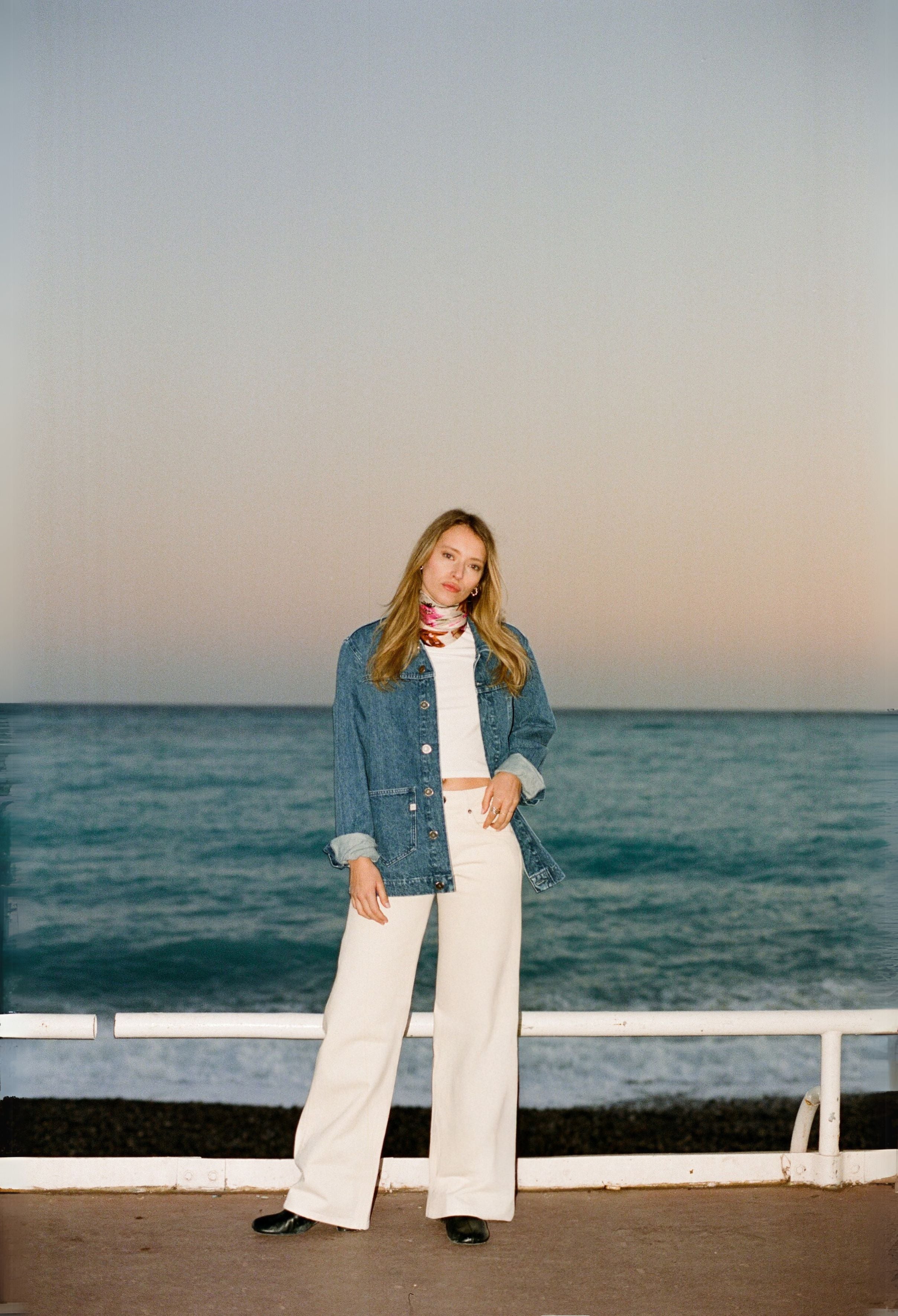 Stylish woman in denim jacket and white pants, standing on a boardwalk with pastel sky and ocean backdrop, wearing a scarf.