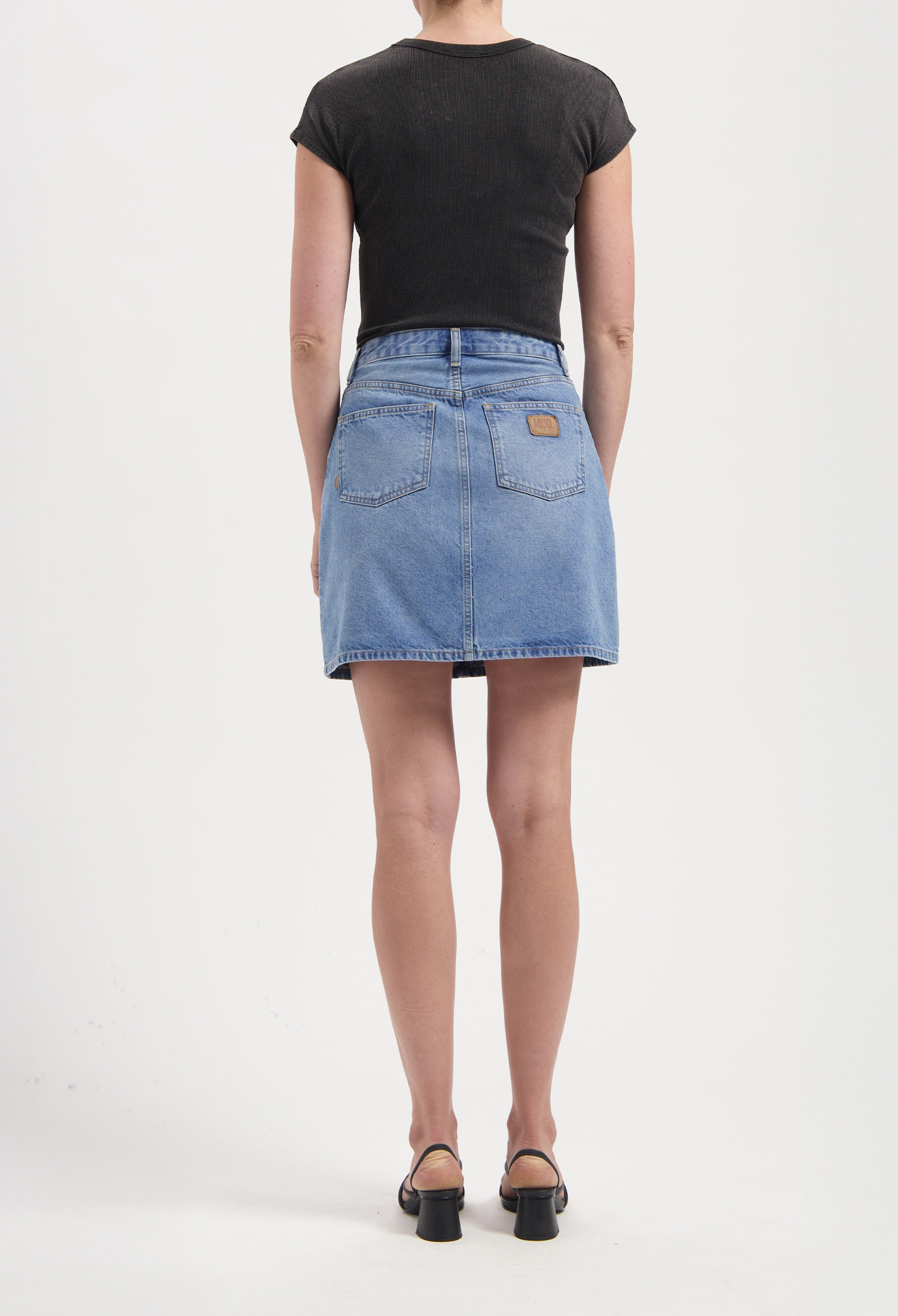Back view of person wearing a blue denim skirt and black short-sleeve top paired with black heeled sandals.