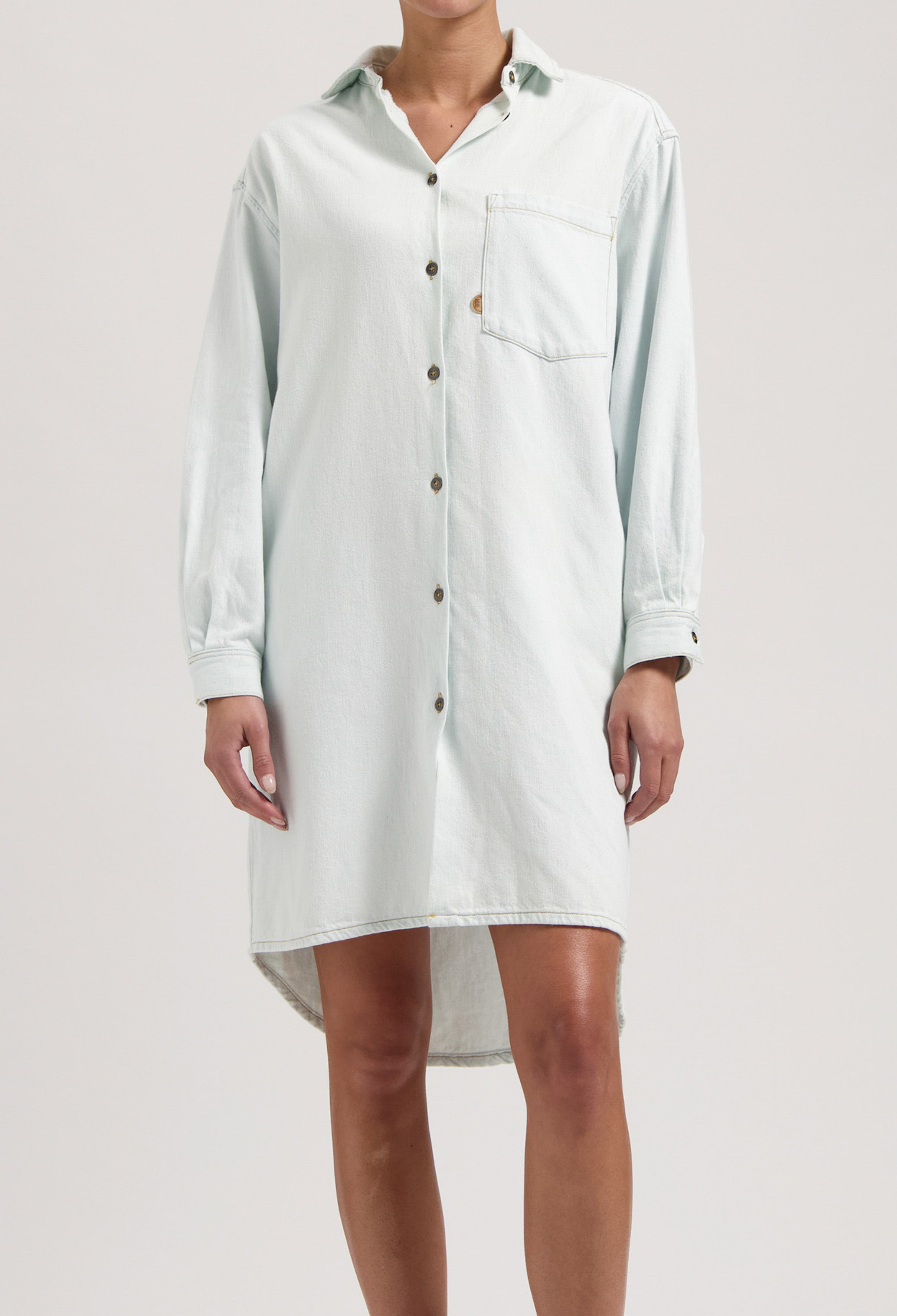 Loose-fitting white denim shirt dress with button front, featuring a single front pocket and long sleeves, worn casually.