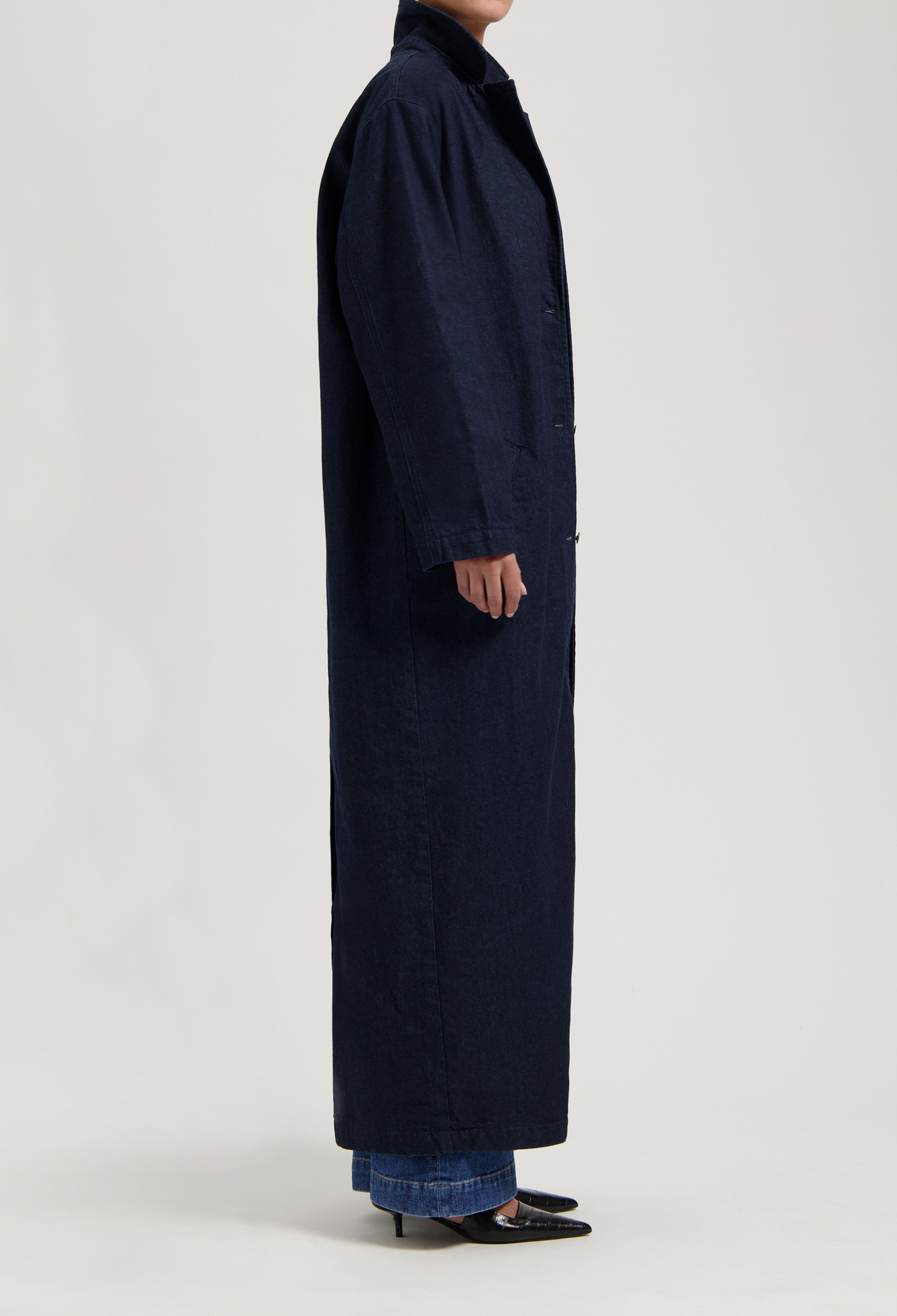 Long navy trench coat in dark fabric, styled with blue denim jeans and black pointed heels, side view.