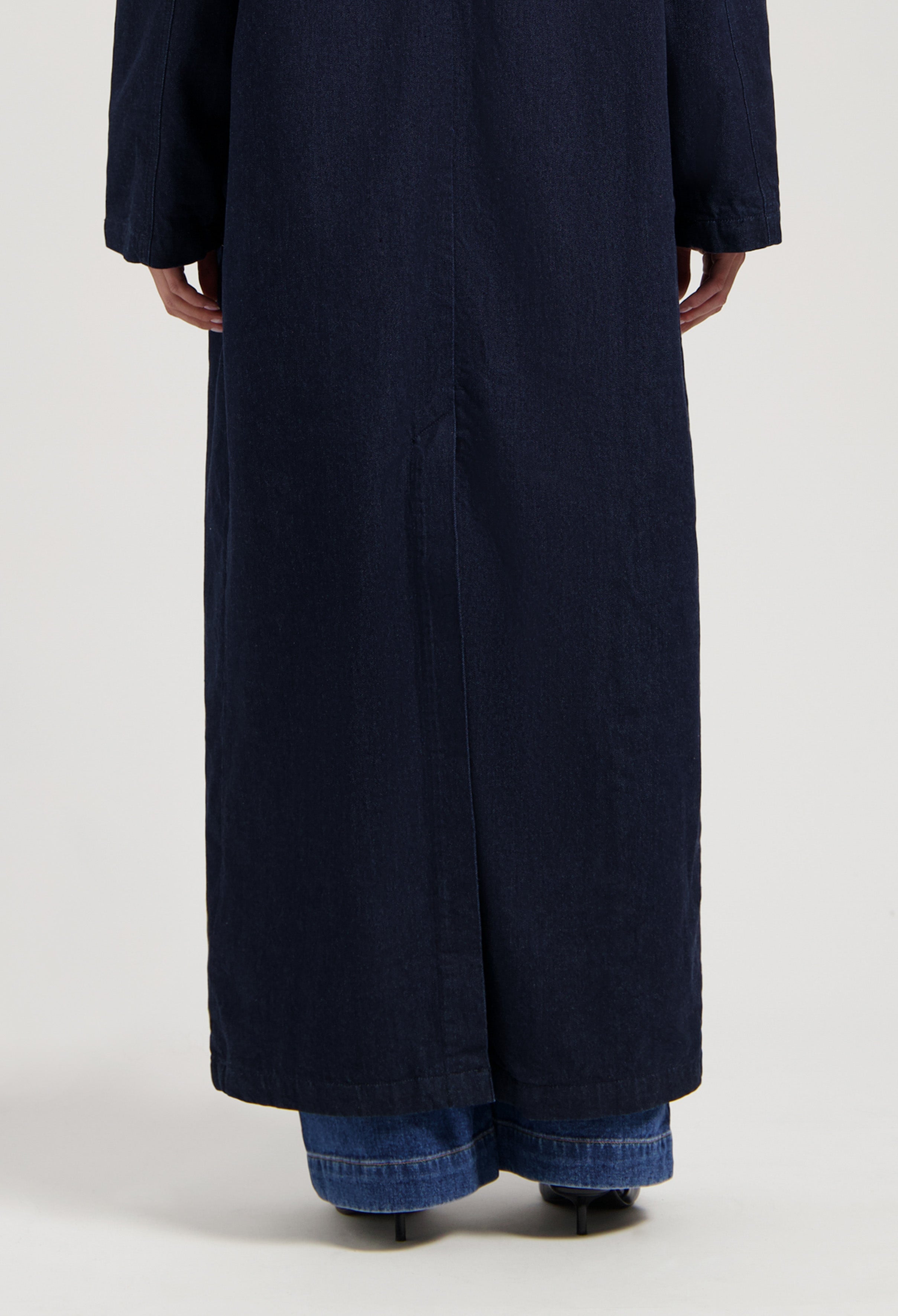 Dark blue denim trench coat shown from the back, styled over blue jeans and black heeled boots.