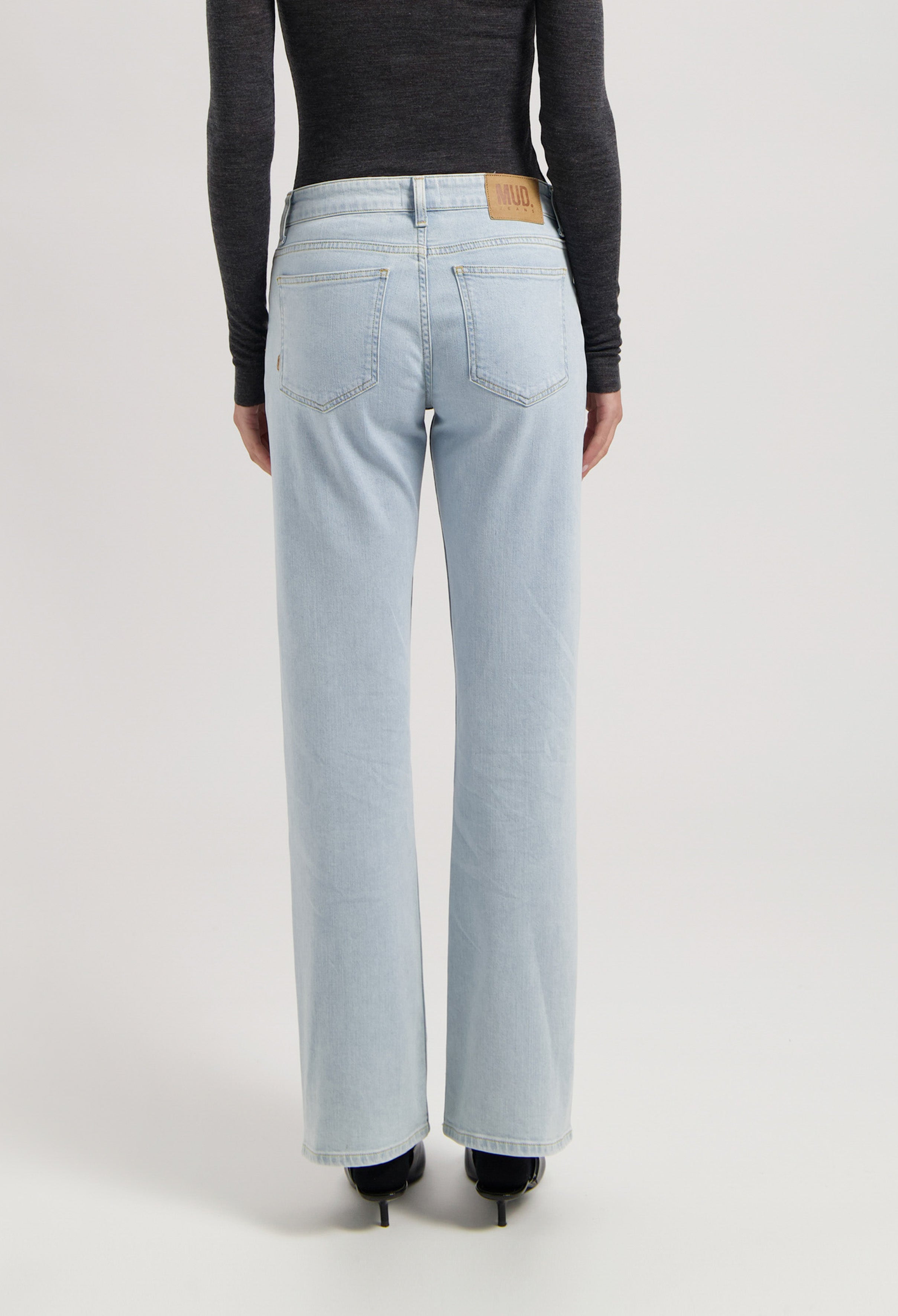 Back view of light blue, straight-leg jeans from MUD Jeans, paired with a dark gray long-sleeve top and black heels.