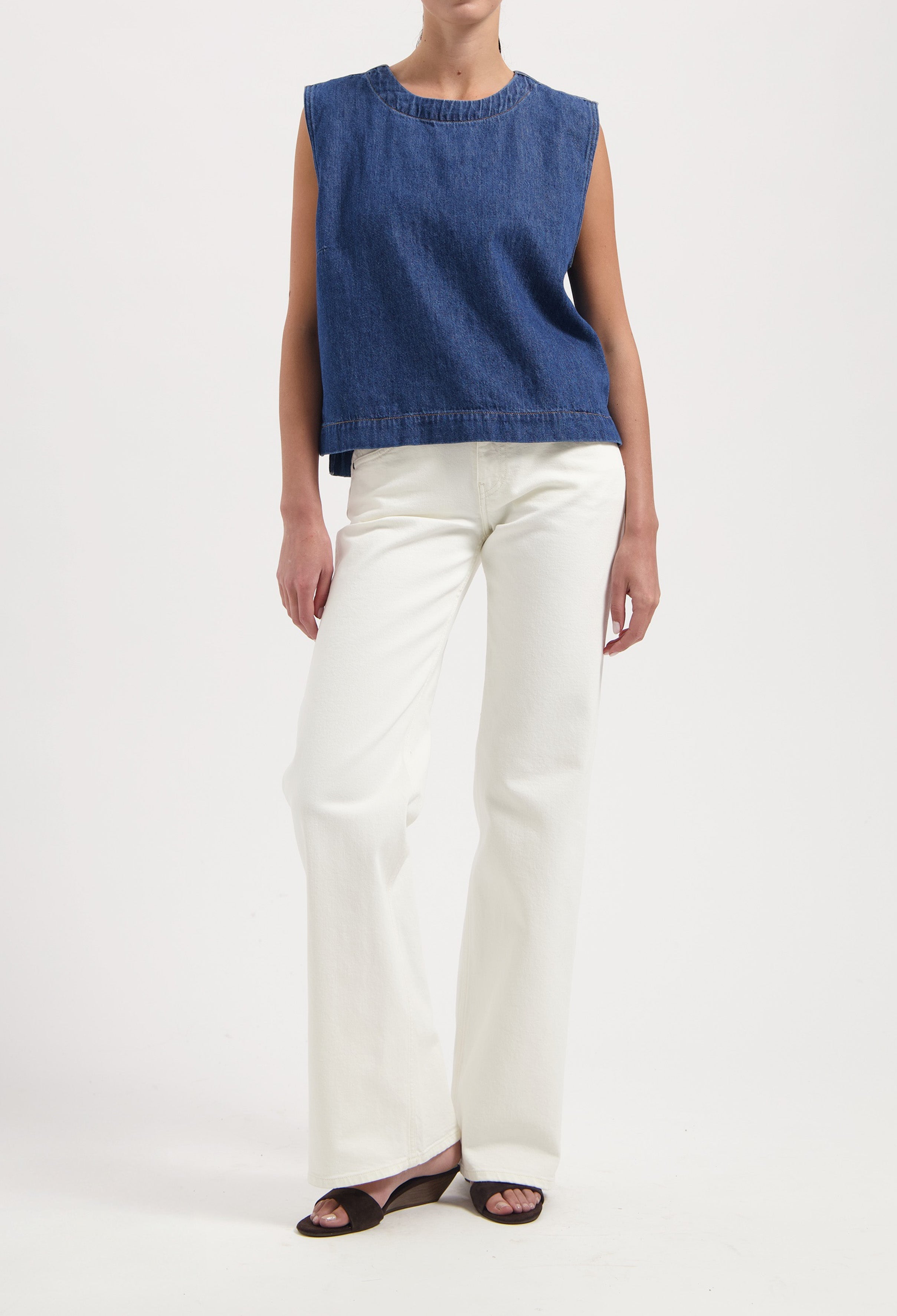 Woman wearing a sleeveless blue denim top and white wide-leg pants, styled with black open-toe sandals.