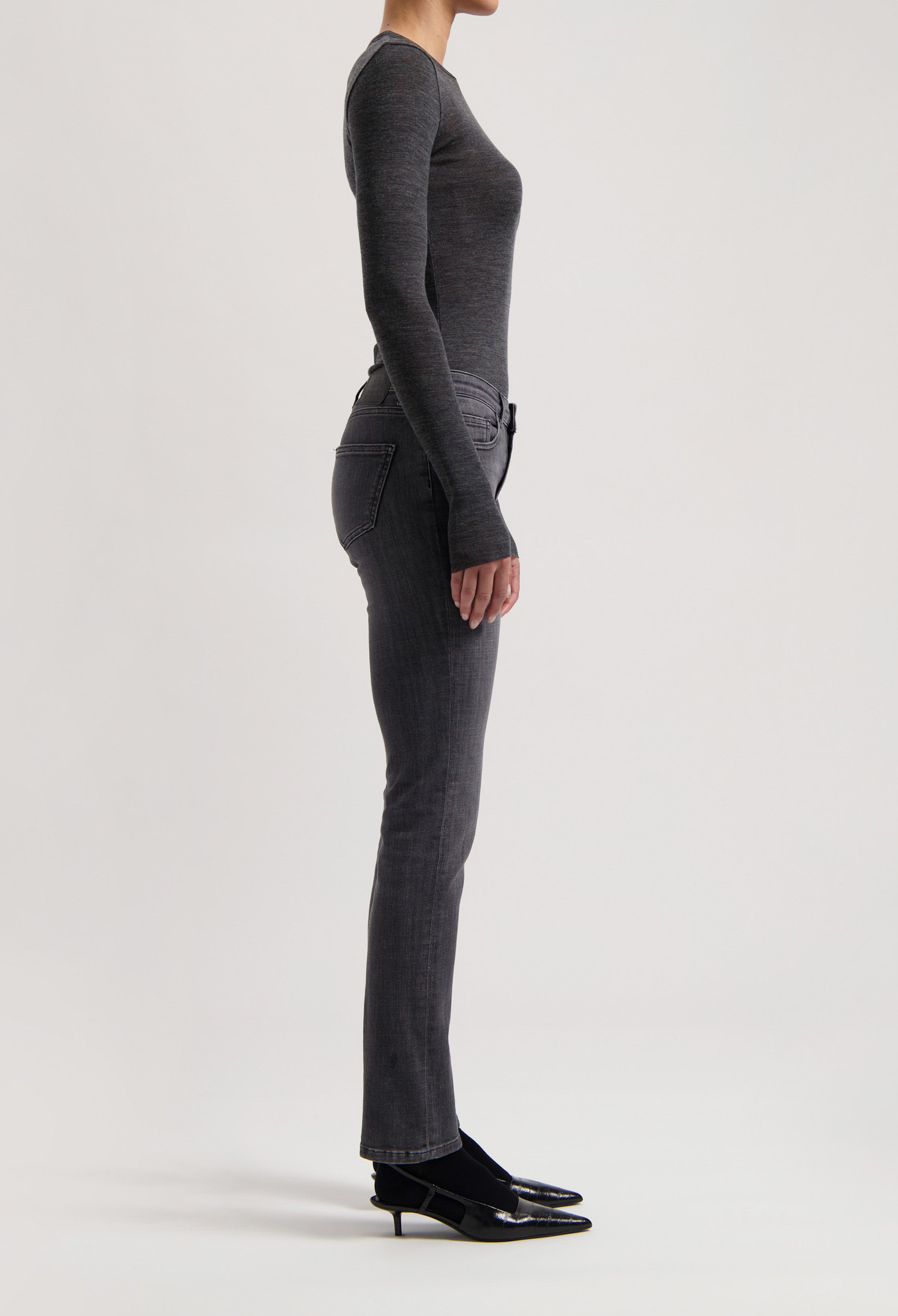 Model wearing a fitted, long-sleeve charcoal top and slim-fit gray jeans with black heels, side profile view.