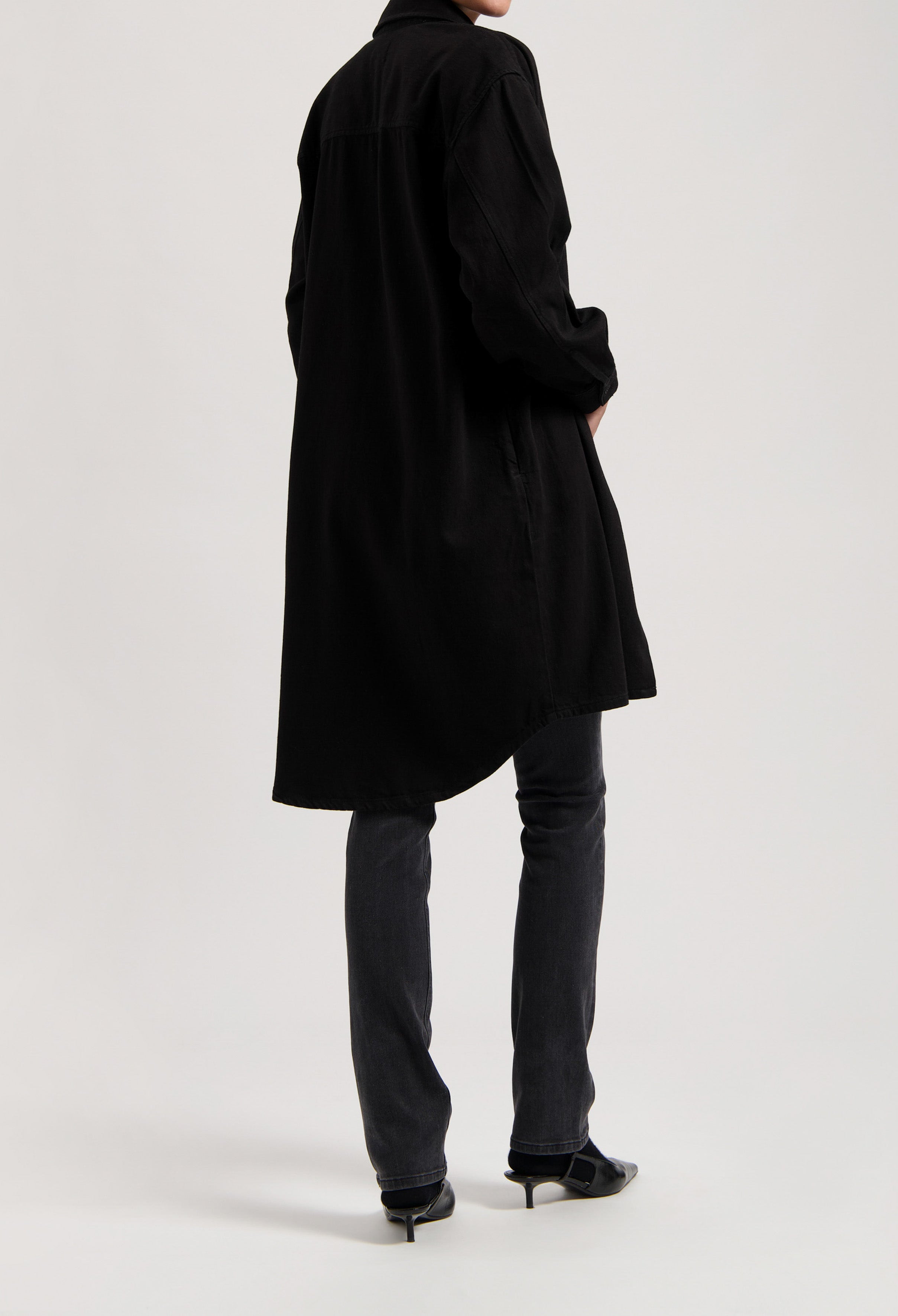 Long black shirt dress styled over dark denim jeans and black pointed-toe heels, showcasing a minimalist fashion look.