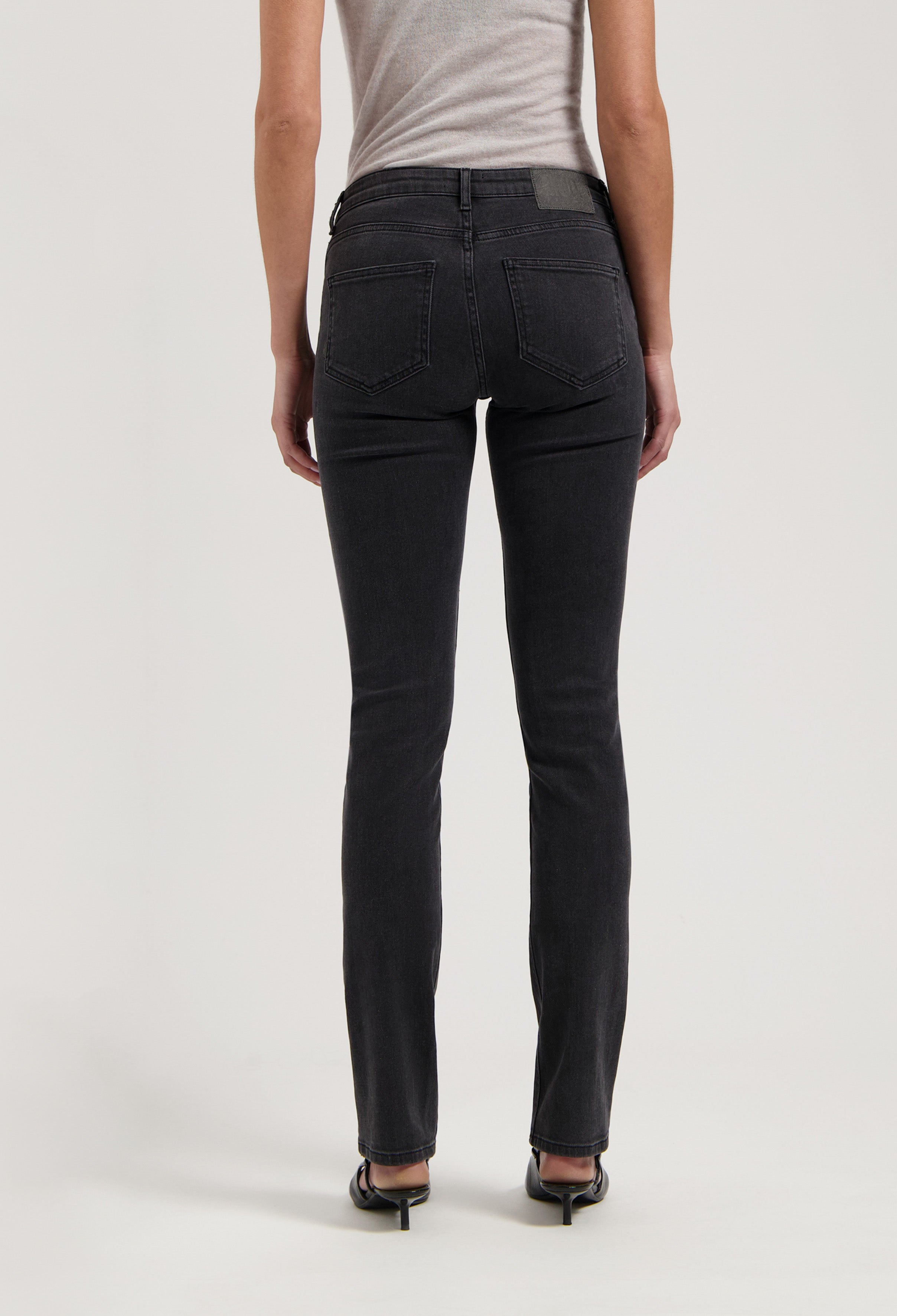 Dark gray slim-fit jeans on a model, highlighting the back pockets and straight leg cut, paired with a simple gray top.