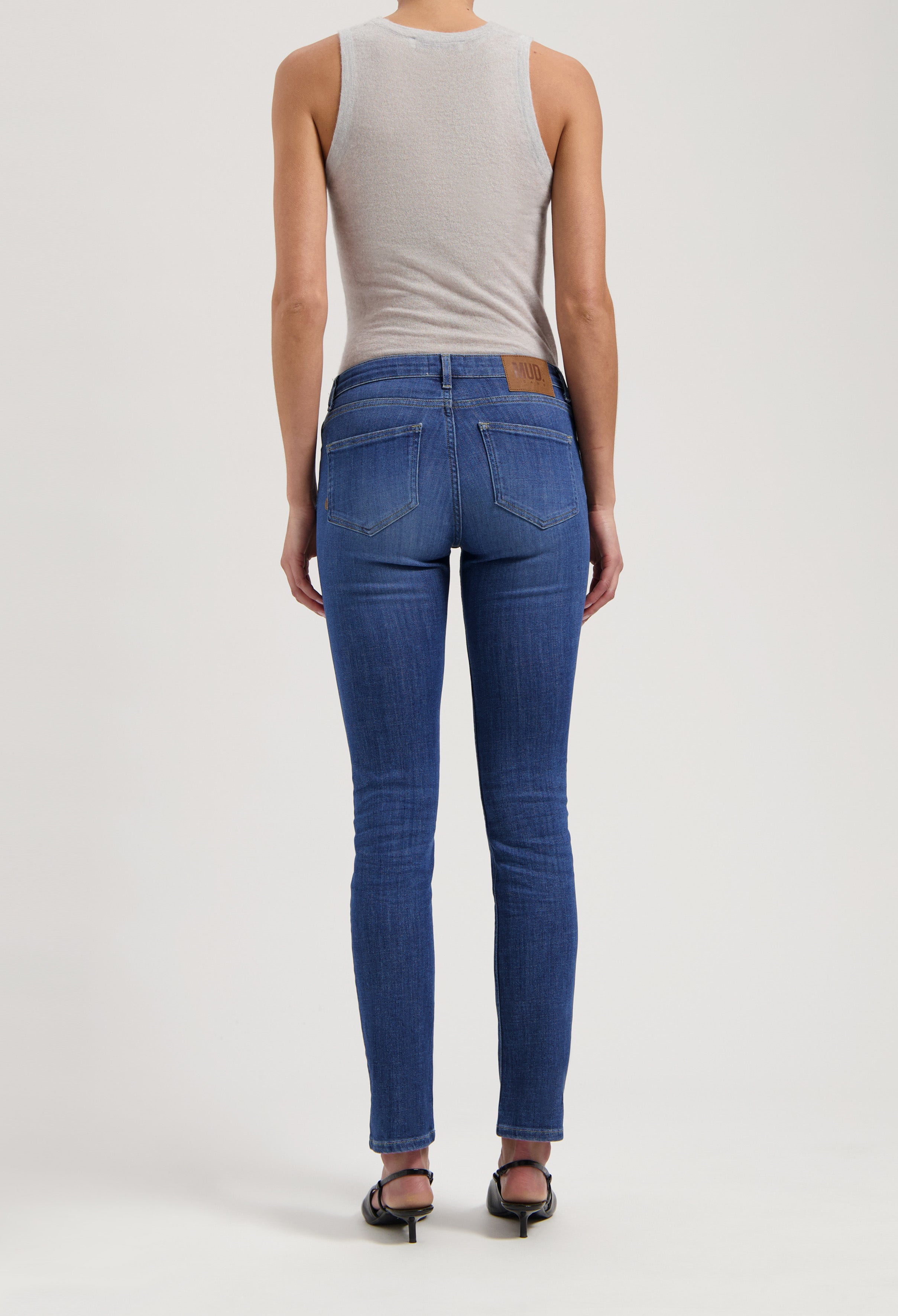 Back view of slim-fit, sustainable blue jeans by MUD Jeans, paired with a light gray sleeveless top and black sandals.