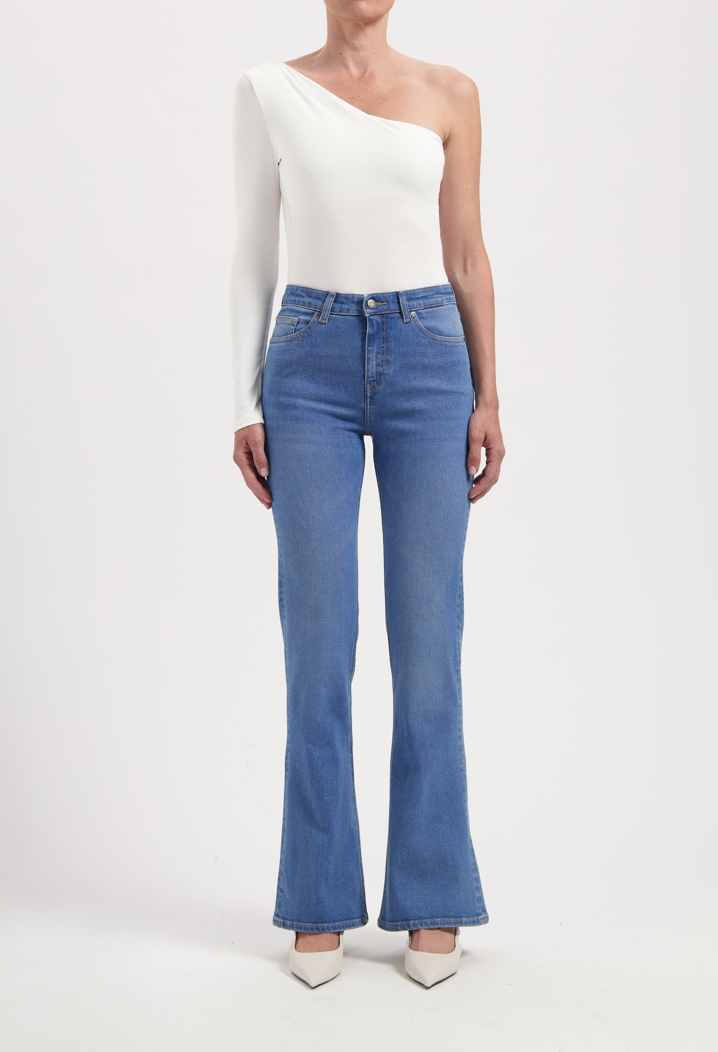 One-shoulder white top with long sleeve paired with high-waisted blue flare jeans, styled with white pointed heels.