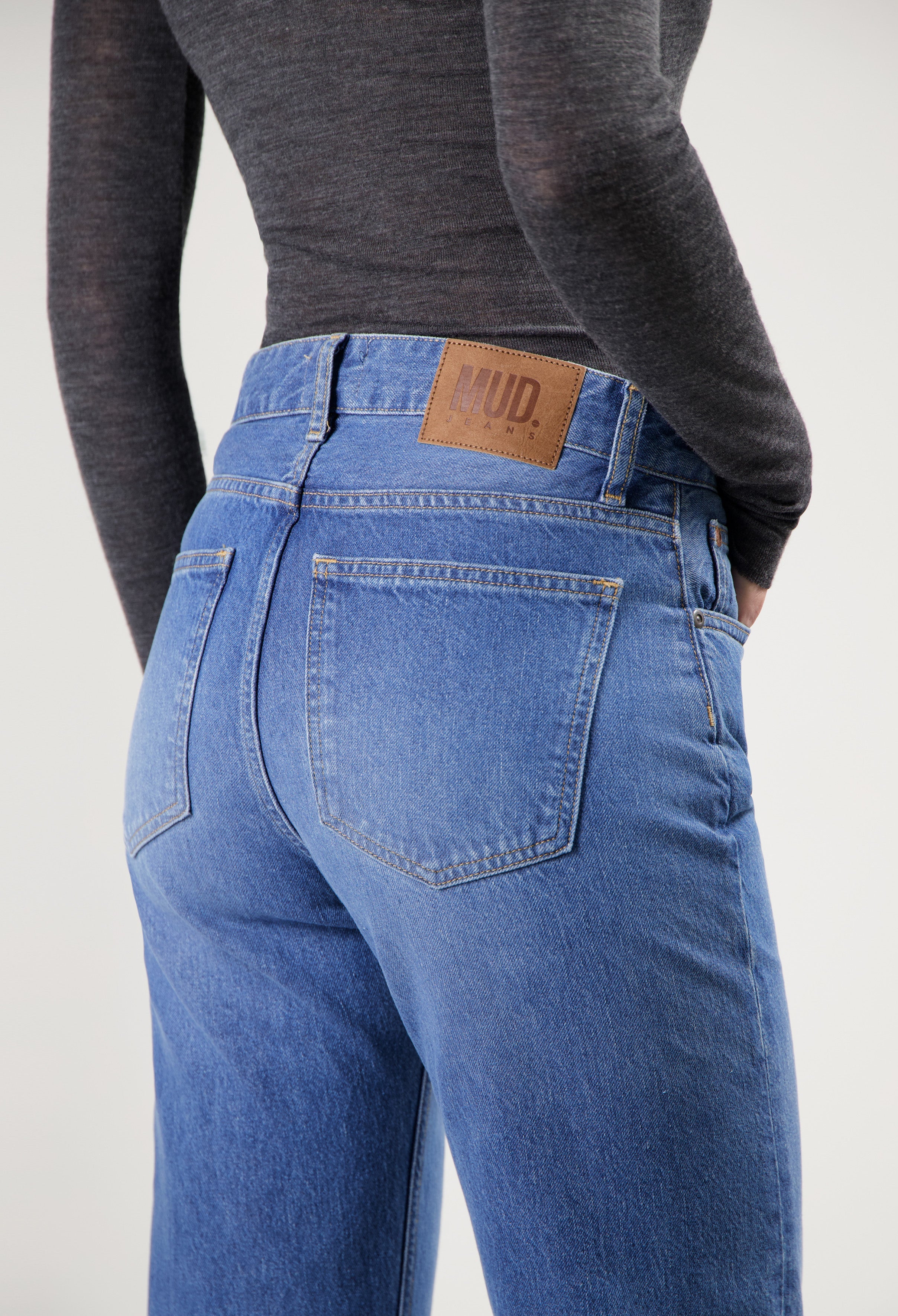 Back view of sustainable MUD Jeans in blue denim, high-waisted fit, paired with a dark gray long-sleeve top.