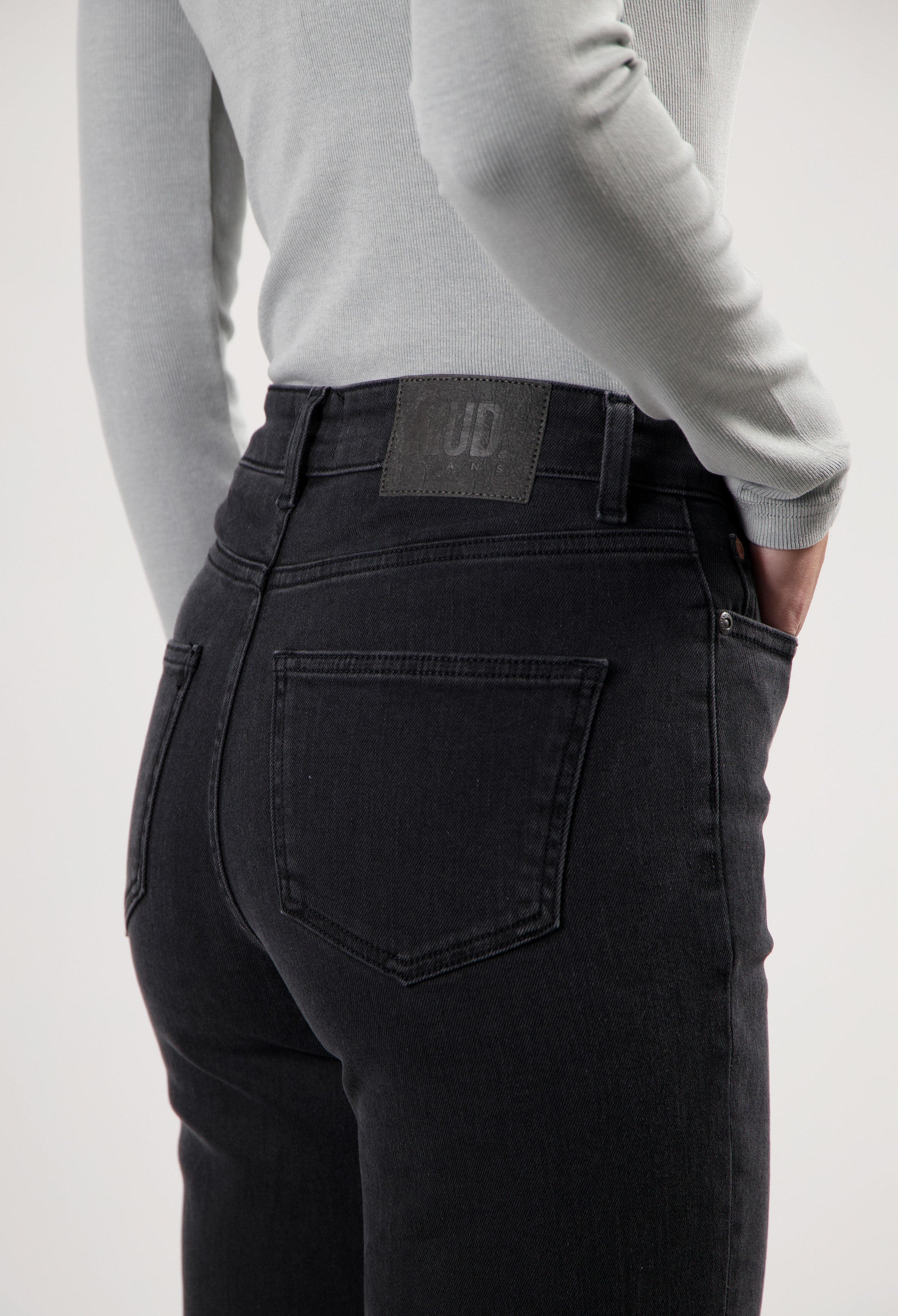 High-waisted, slim-fit, black denim jeans by MUD Jeans, worn with a long sleeve light gray top, showcasing back pocket details.