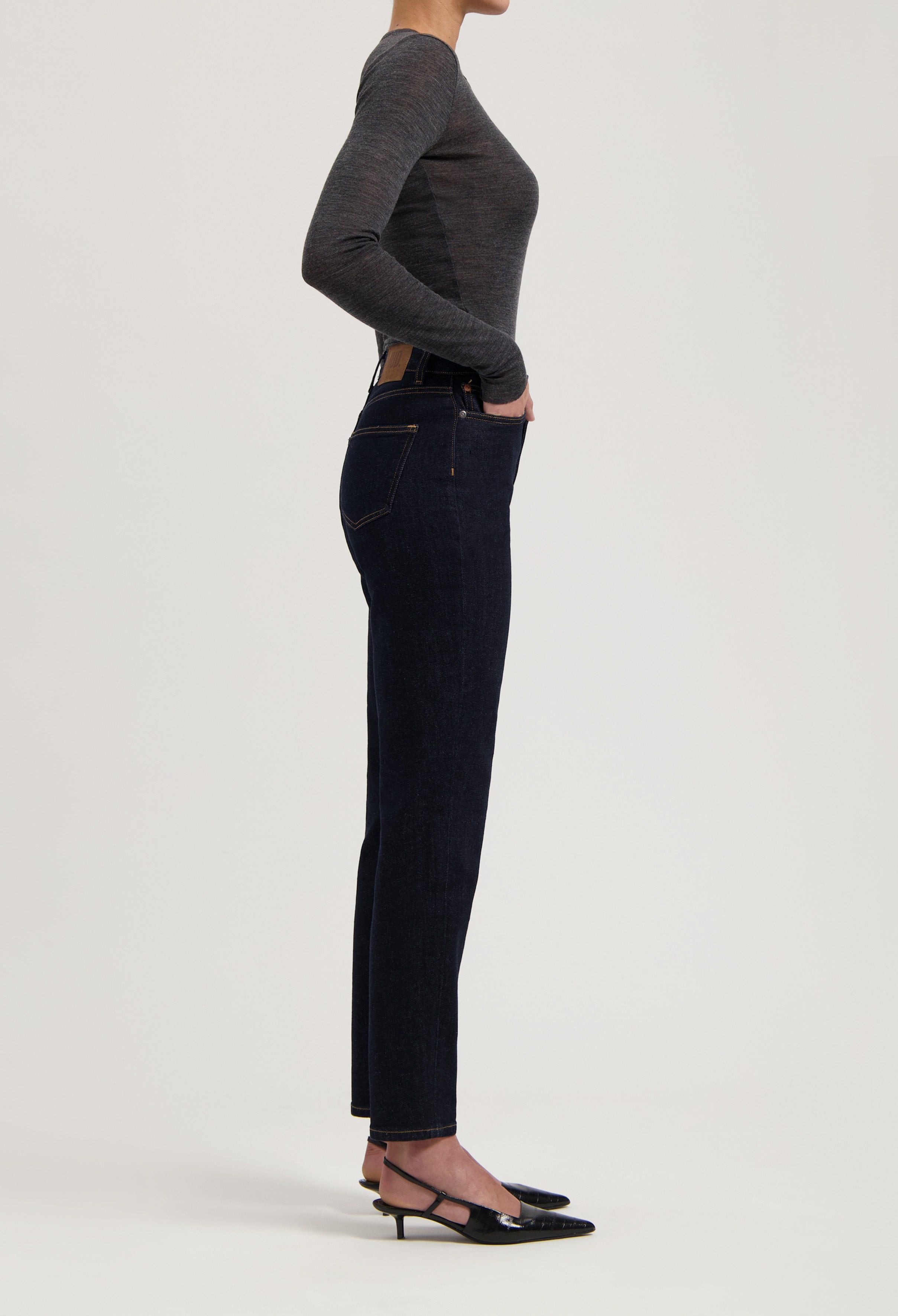 Dark blue, slim-fit denim jeans paired with a gray long-sleeve top, showcasing a side profile and black low-heel shoes.