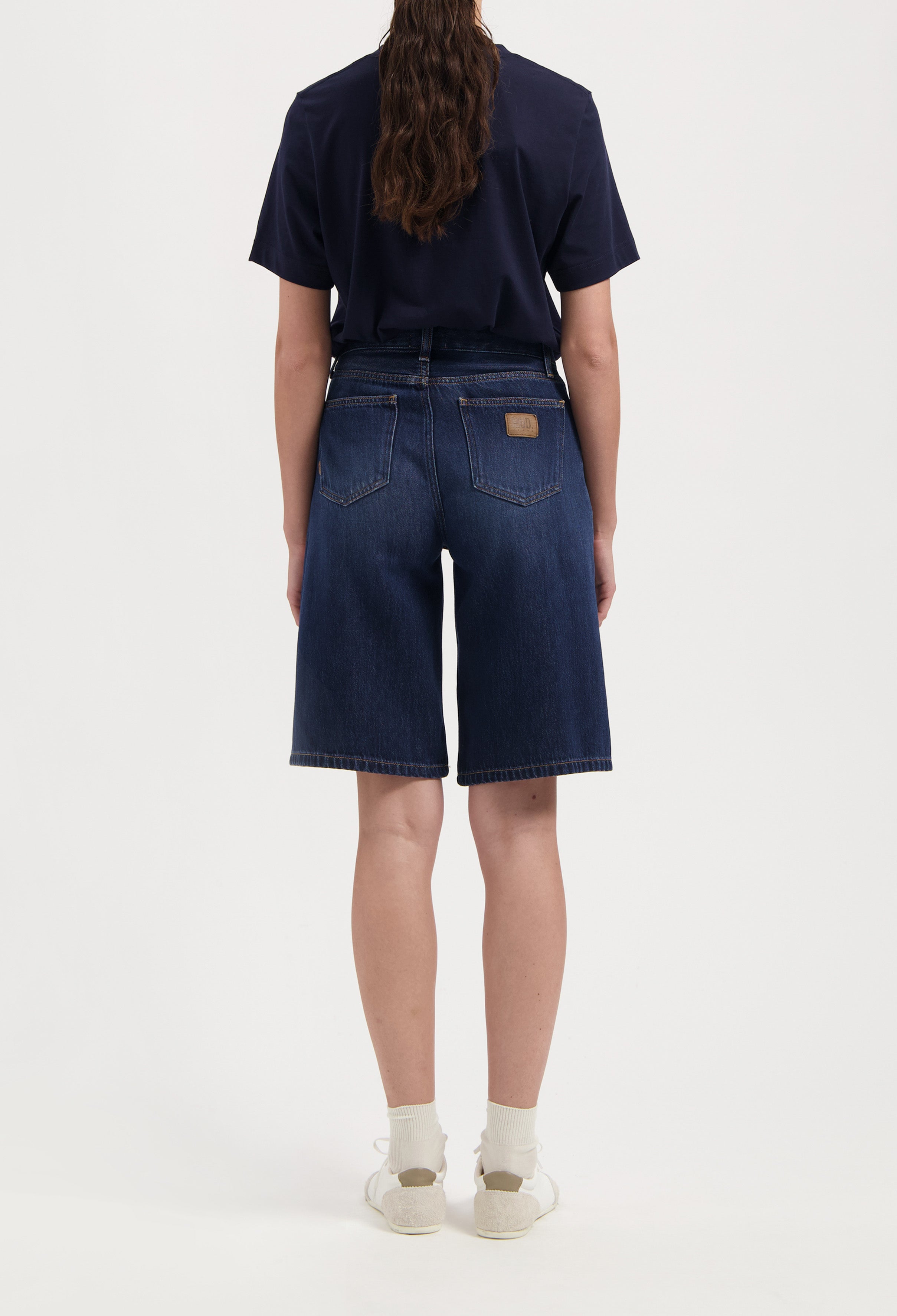 Back view of dark blue denim shorts paired with a black tee, showcasing relaxed fit and classic pocket design with white sneakers.