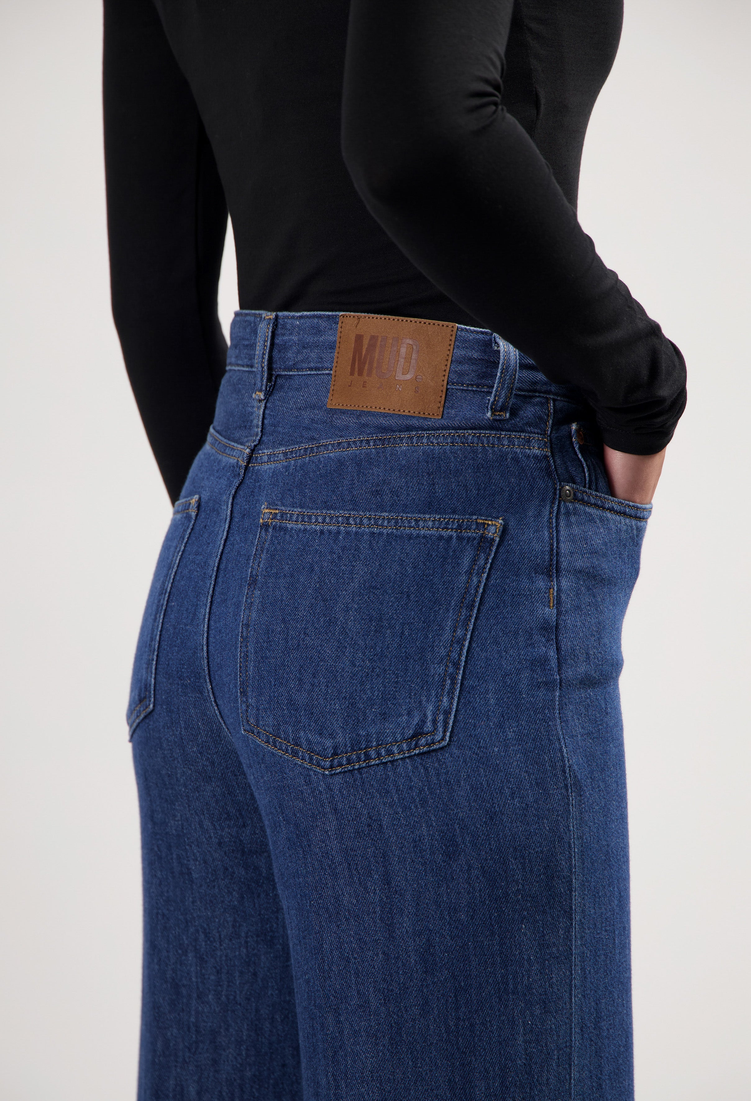 Person wearing blue MUD Jeans, showcasing sustainable high-waisted denim with a leather patch, paired with a black top.