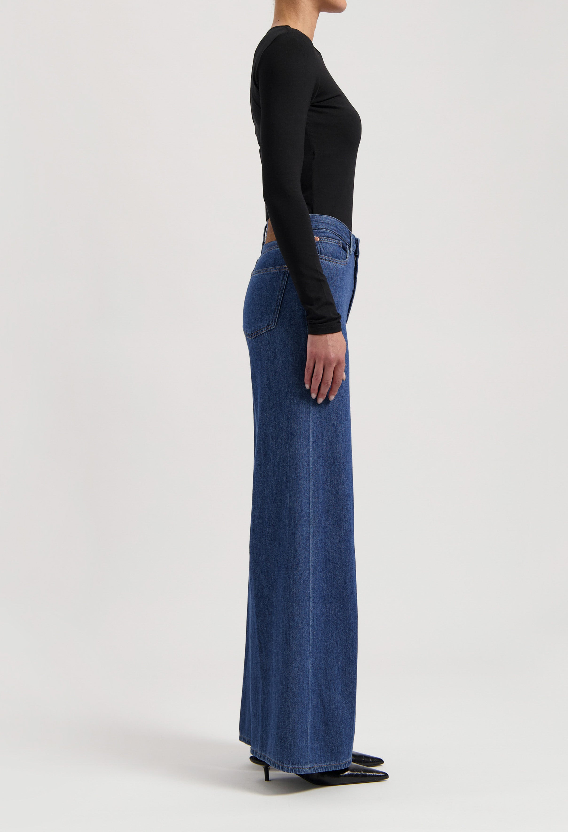 Woman in black long-sleeve top and wide-leg blue denim jeans, side view, standing on white background.
