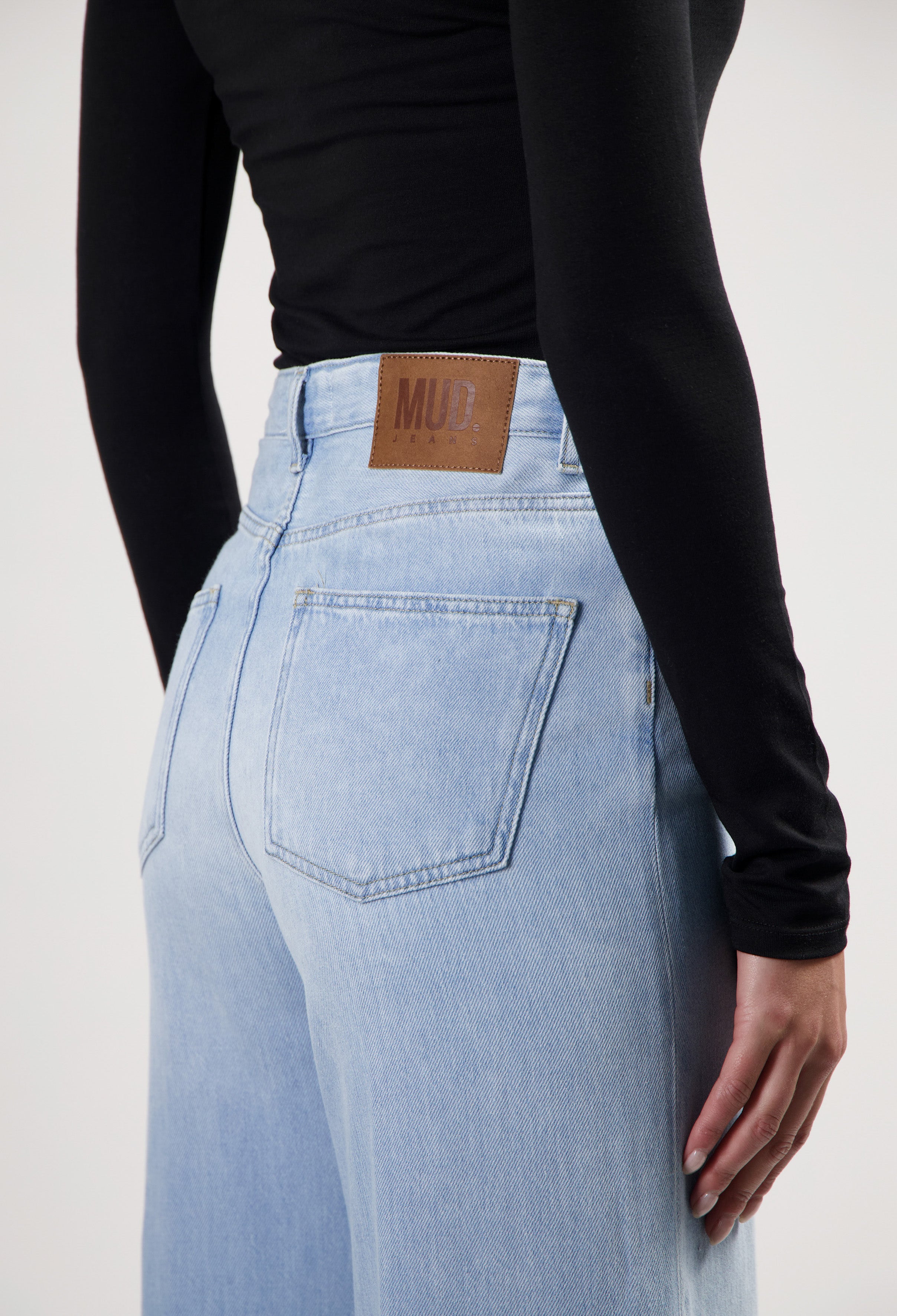 Light blue high-waisted denim jeans by MUD Jeans, featuring wide legs and a vegan leather patch, paired with a black top.