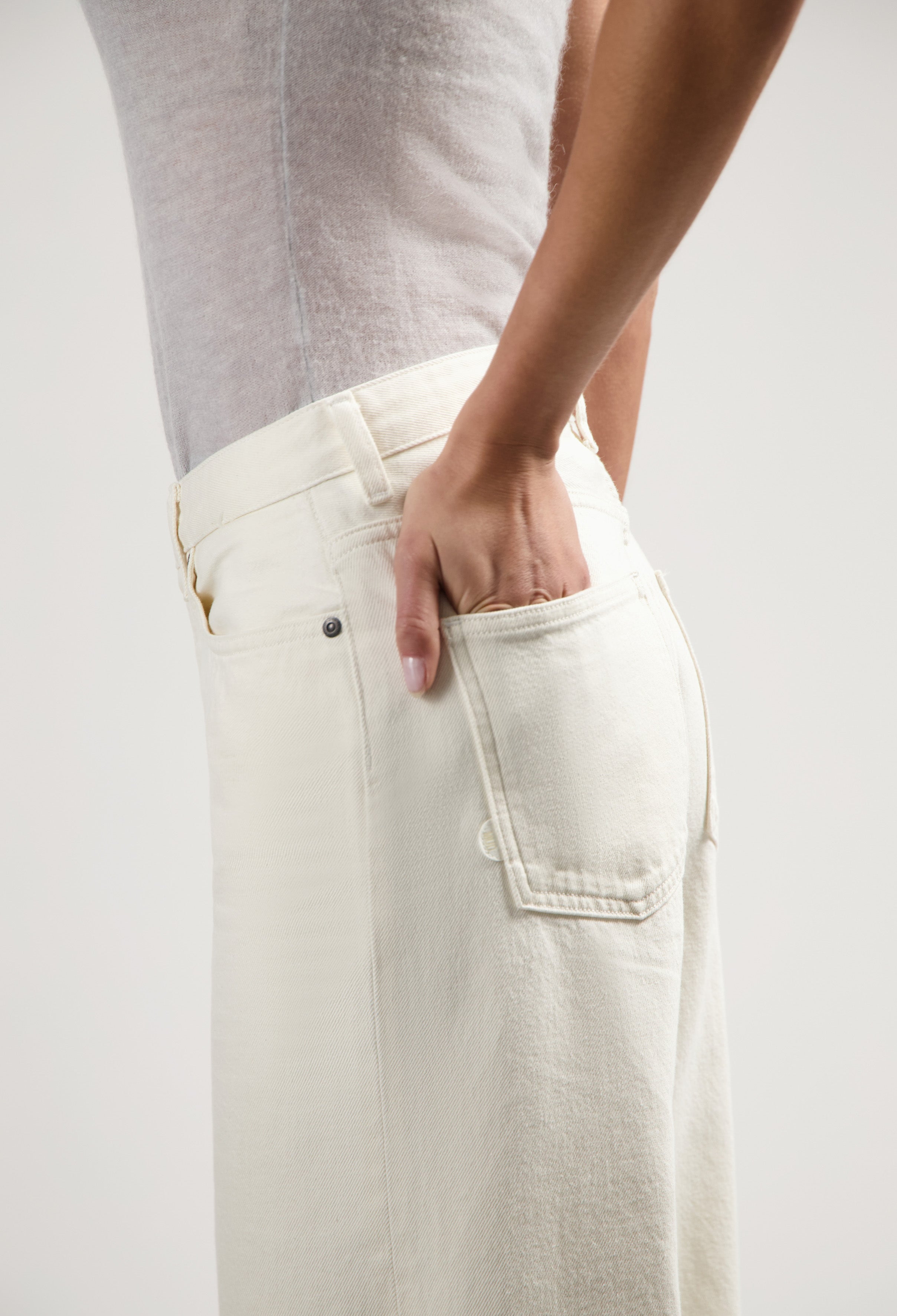 Person wearing off-white high-waisted jeans with a fitted gray sleeveless top, hand in back pocket, side view.