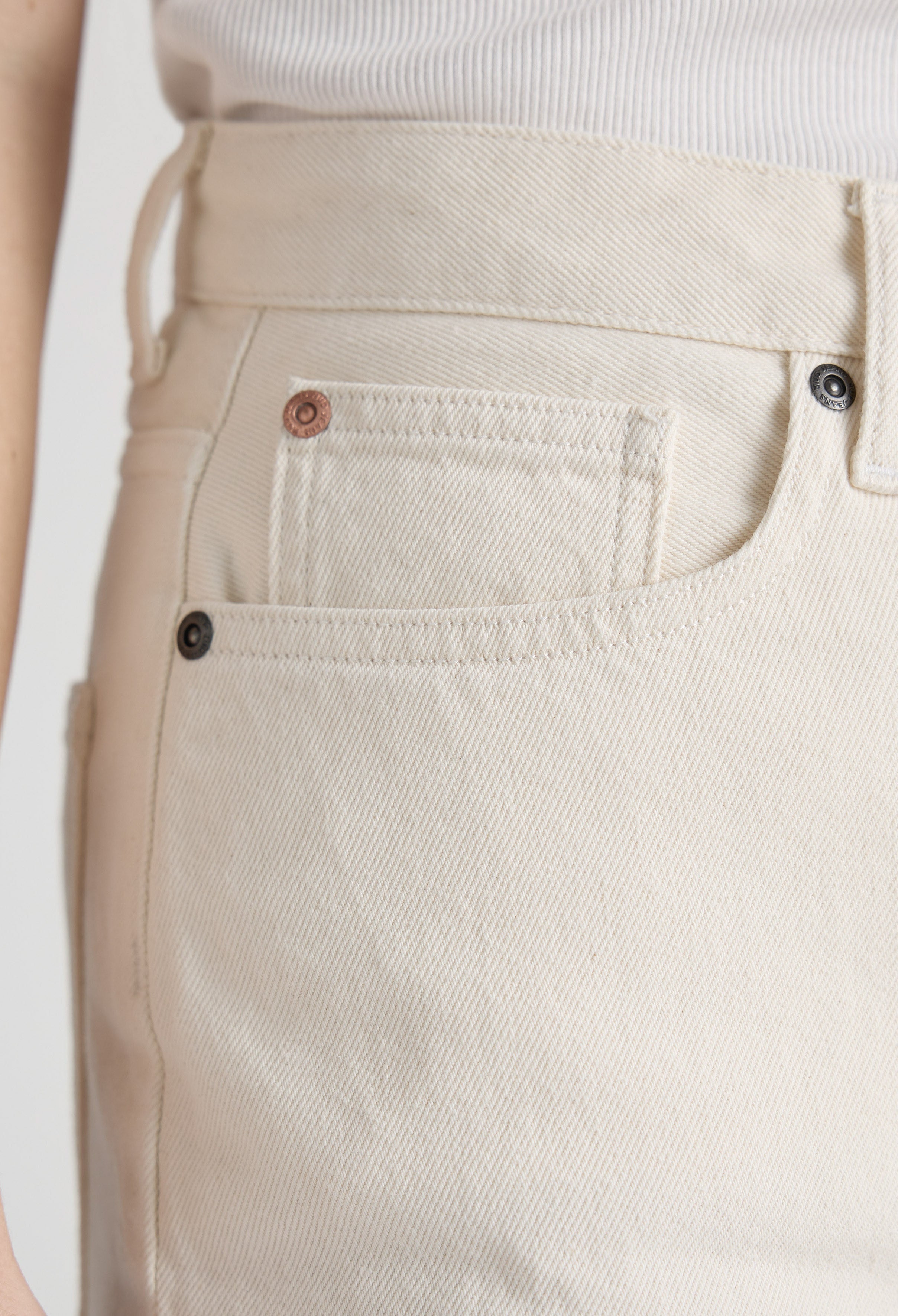 Close-up of cream-colored denim jeans pocket on a textured fabric, showing intricate stitching details and metal rivet buttons.