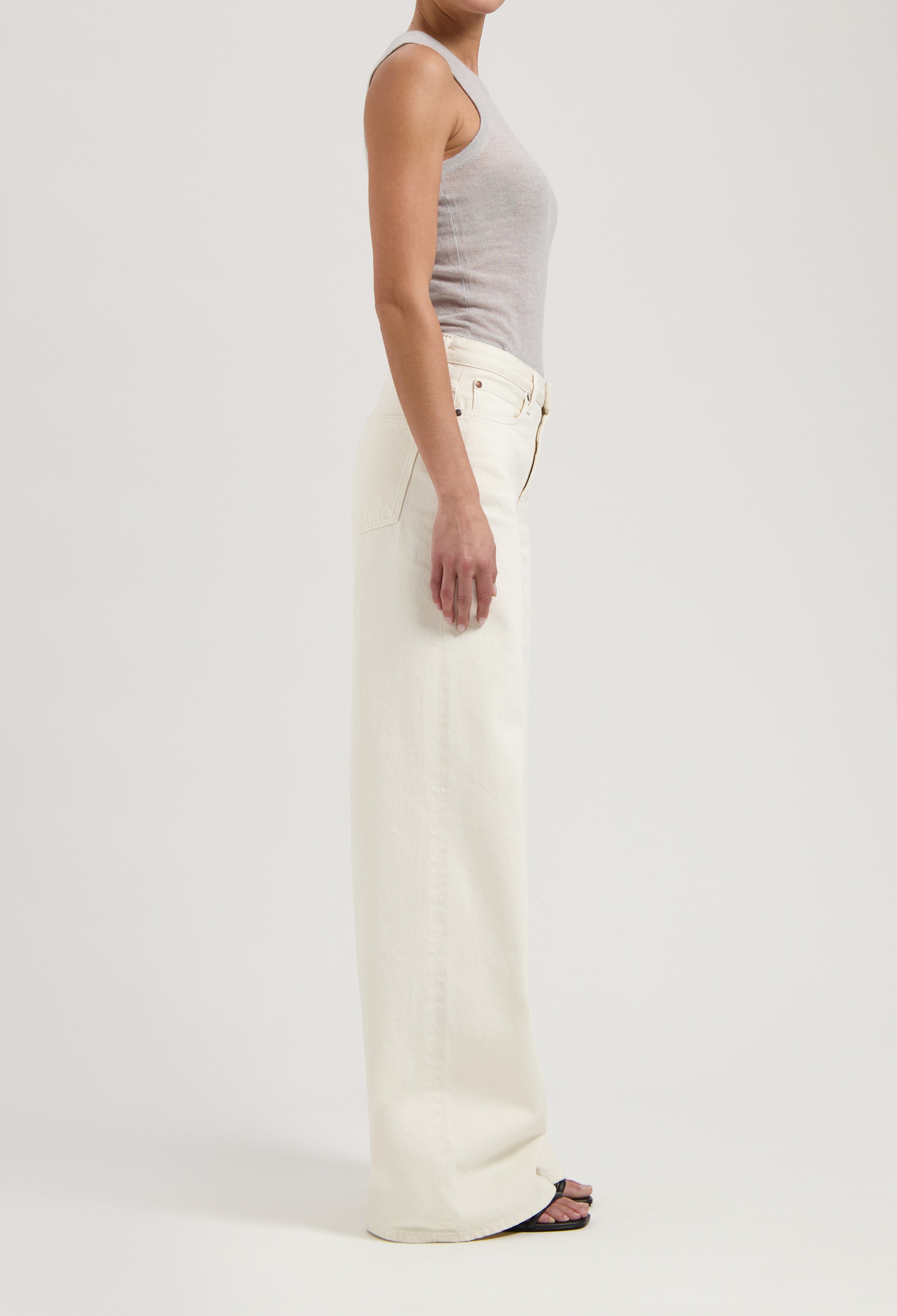A model wearing high-waisted, wide-leg white denim jeans paired with a fitted gray tank top, showcasing a sleek side profile.