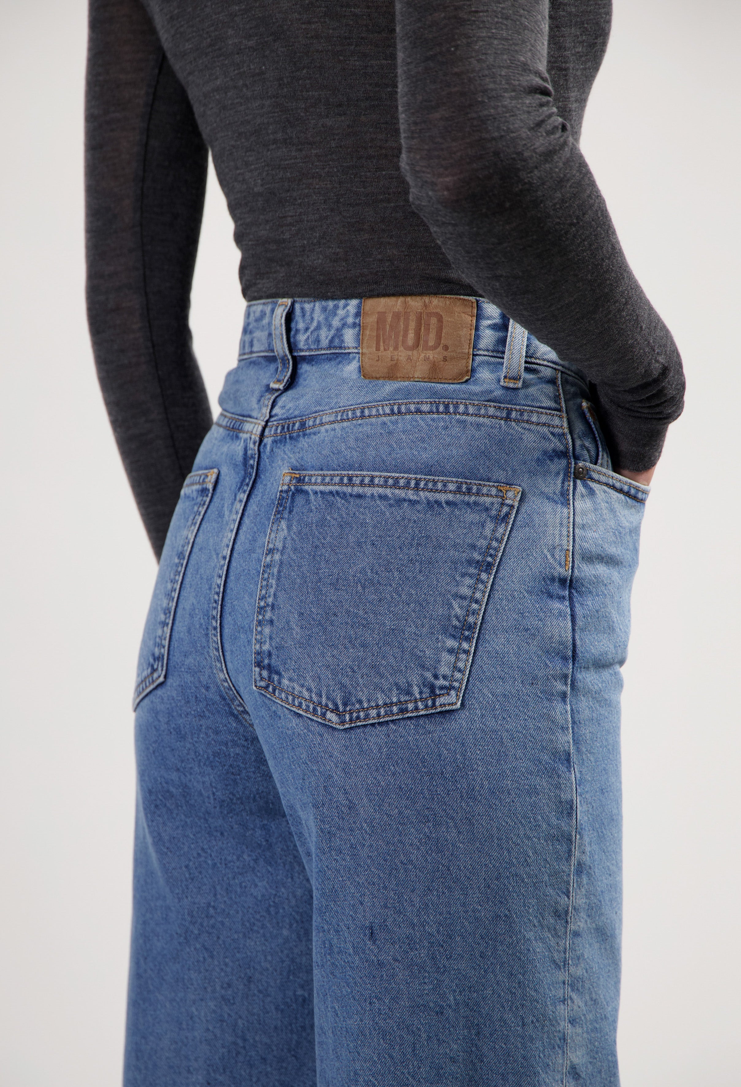 Close-up of MUD Jeans featuring blue denim, high waist design, worn with a form-fitting dark gray long-sleeve top.