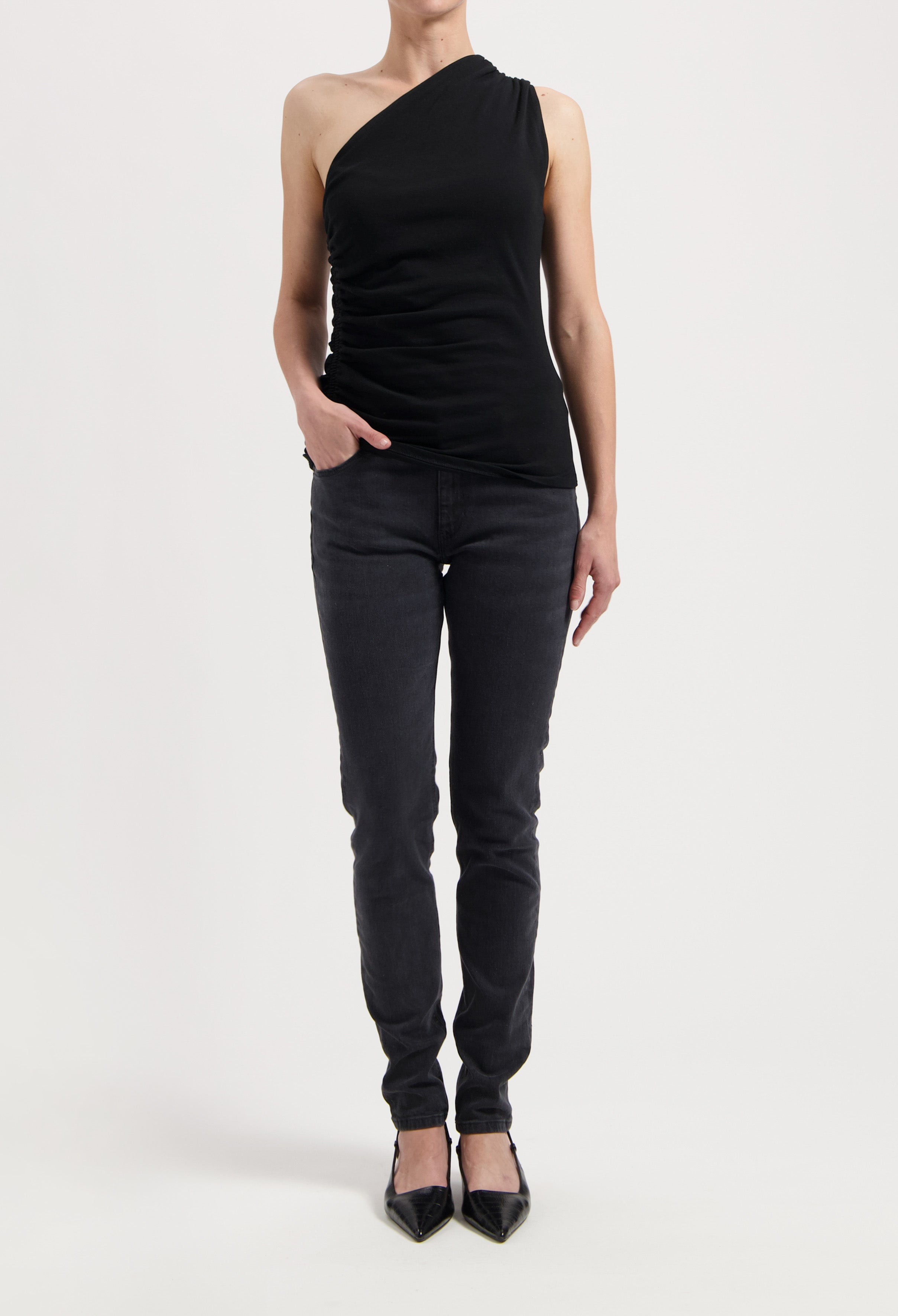 Model wearing an asymmetrical black sleeveless top paired with dark gray slim-fit jeans and black pointed flats.