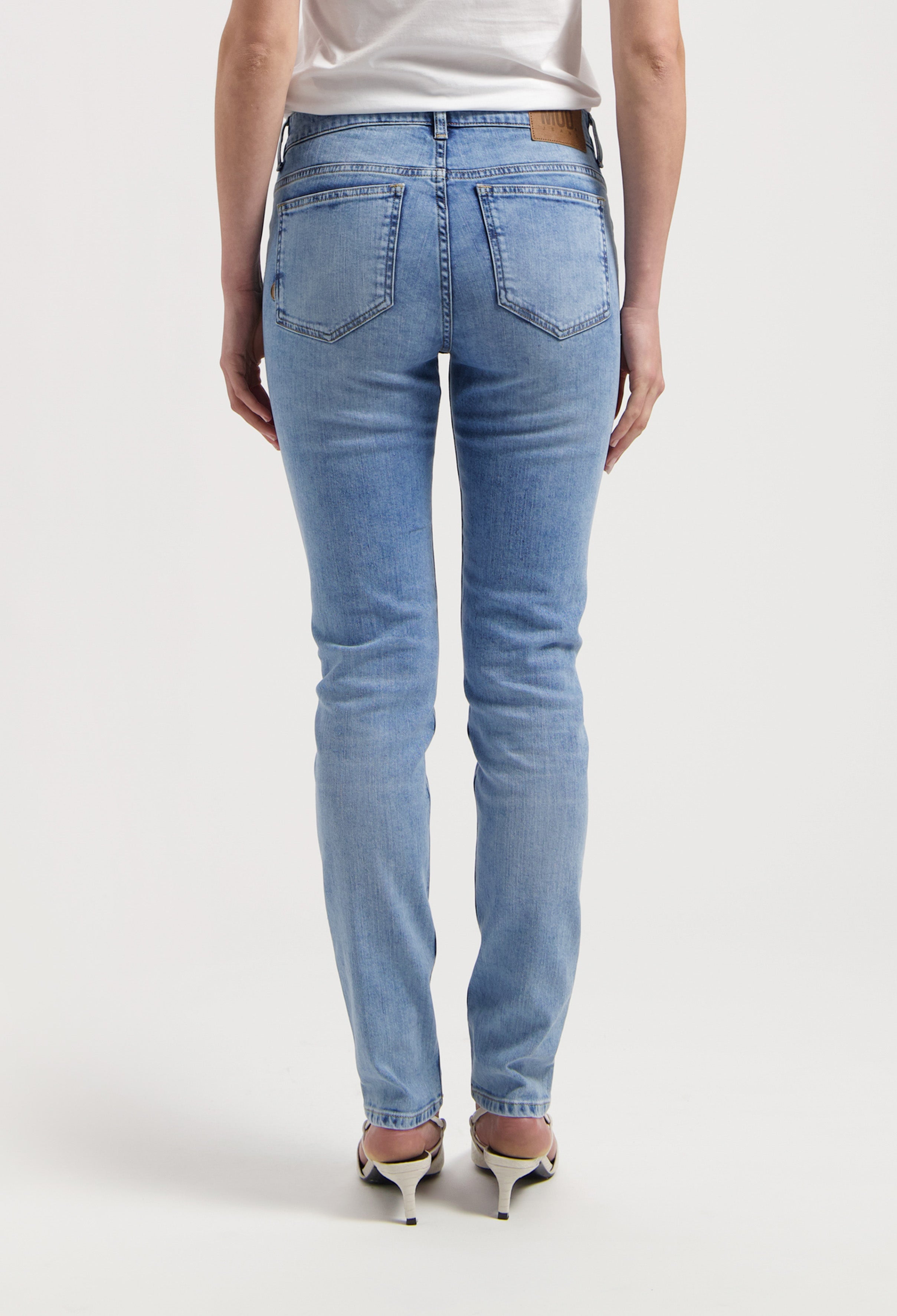 Light blue slim-fit women's jeans from the back, featuring a brown leather patch, paired with beige high heels.