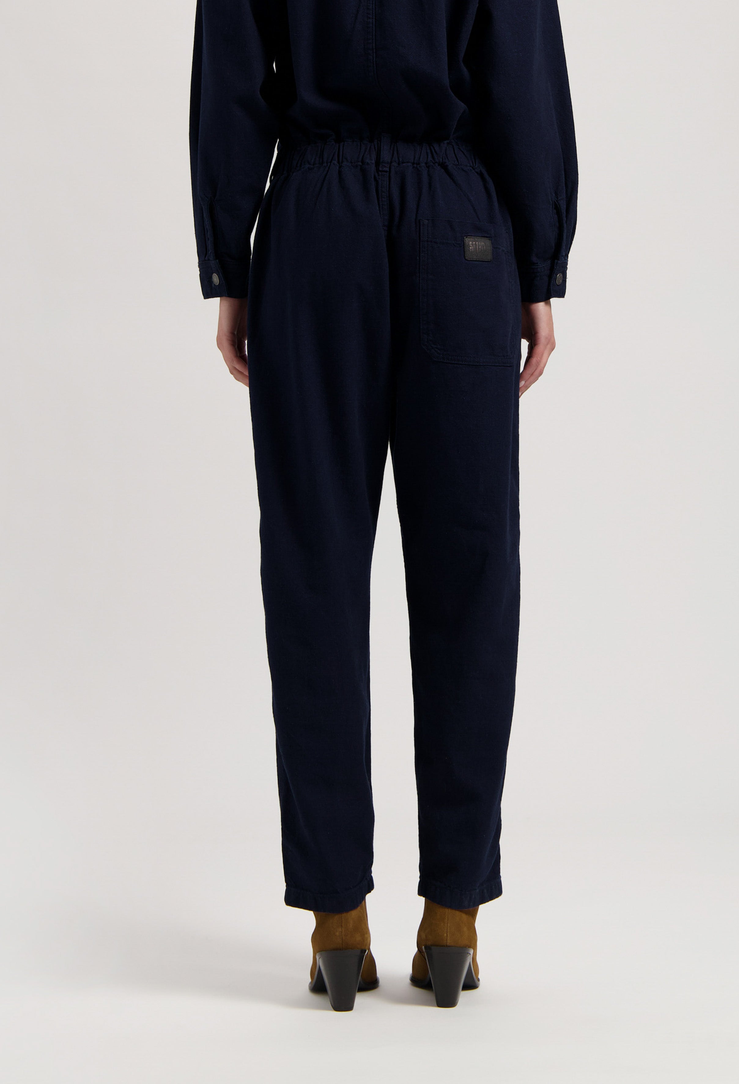 Navy blue cotton jumpsuit with elastic waistband and buttoned cuffs, worn with brown heeled boots viewed from the back.
