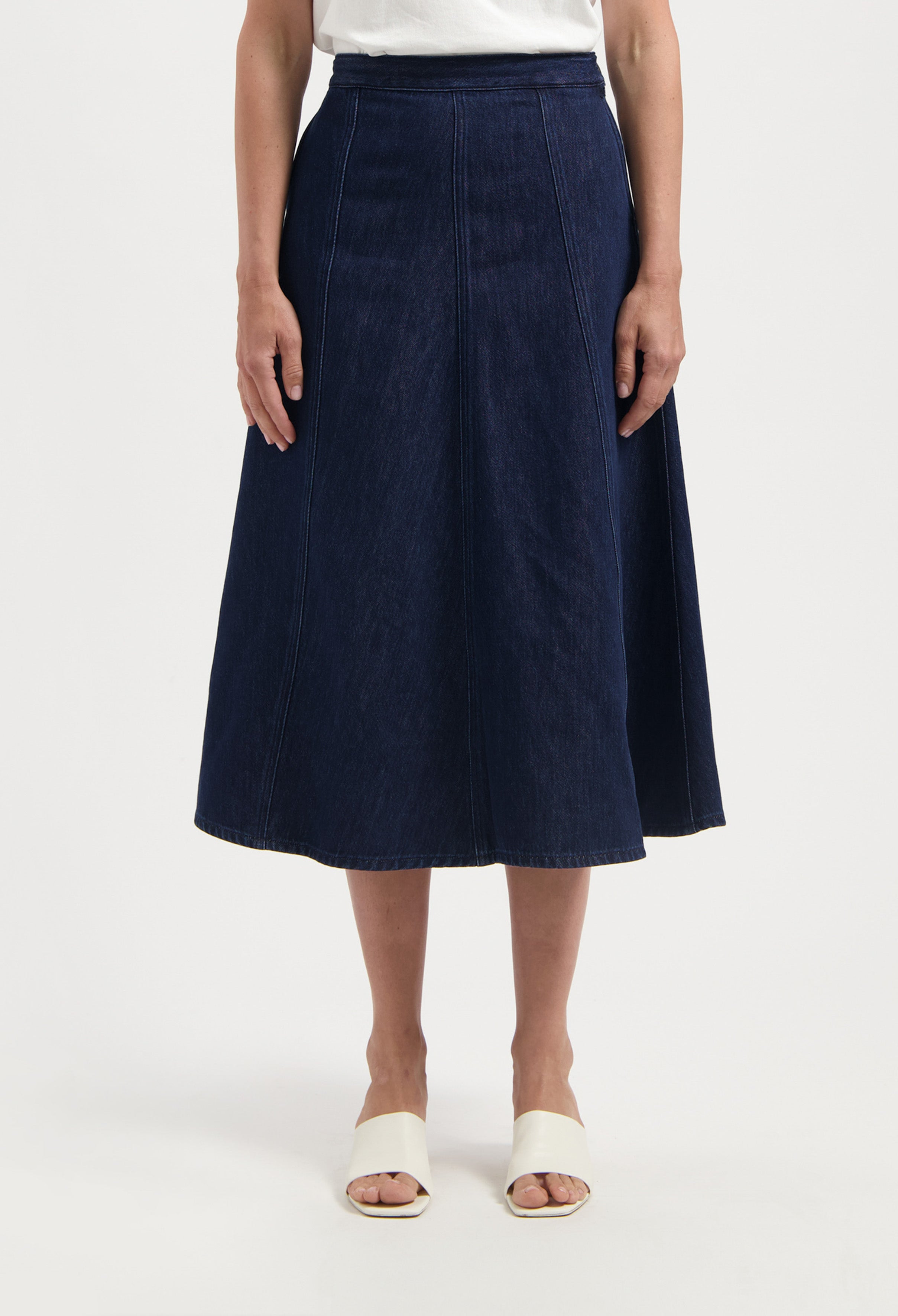 Dark blue, A-line denim skirt worn with a white top, showcasing a classic midi length and vertical seam detailing.
