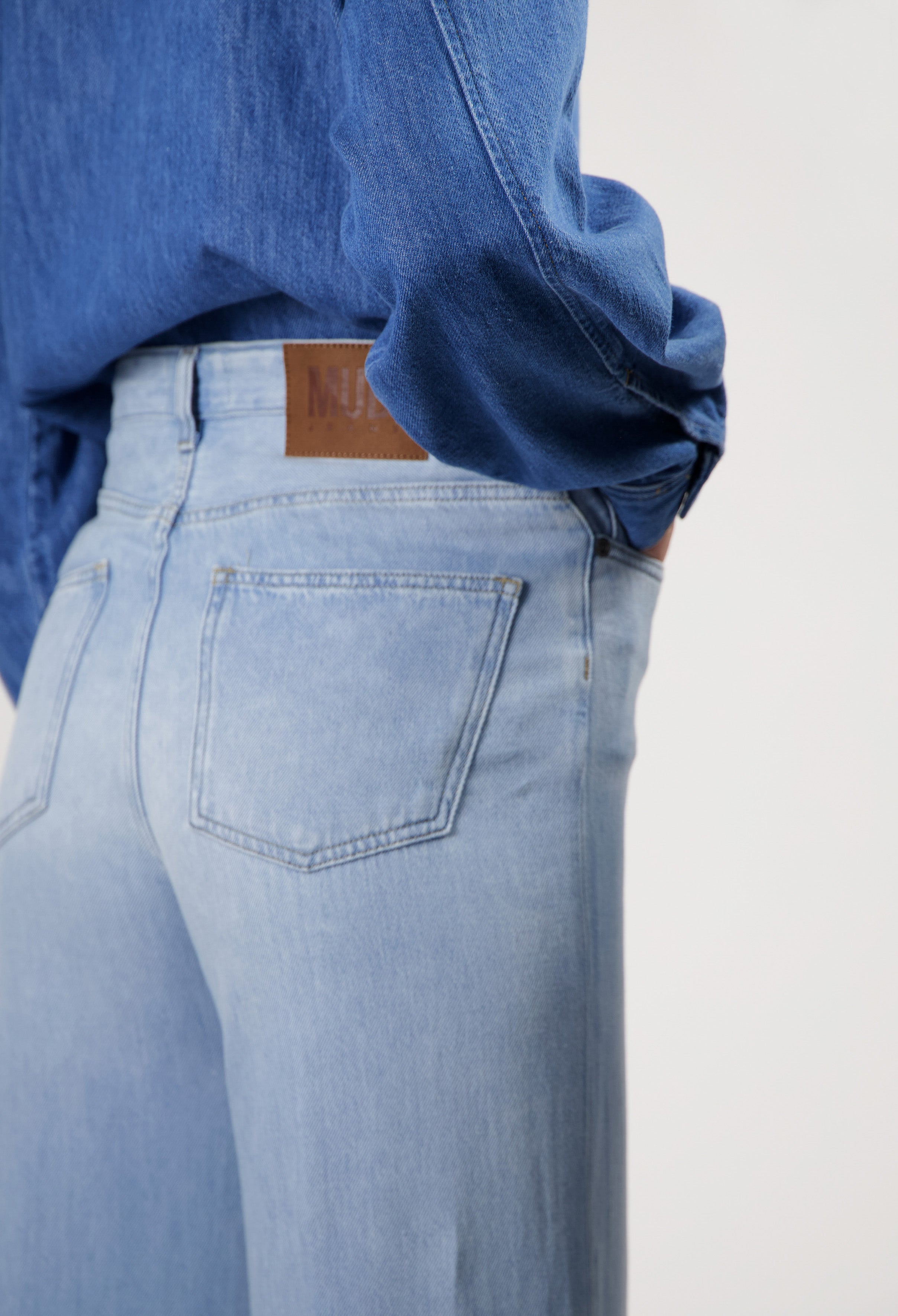 Light blue high-waisted jeans made from organic cotton, featuring classic pocket design and worn with a matching dark blue denim shirt.