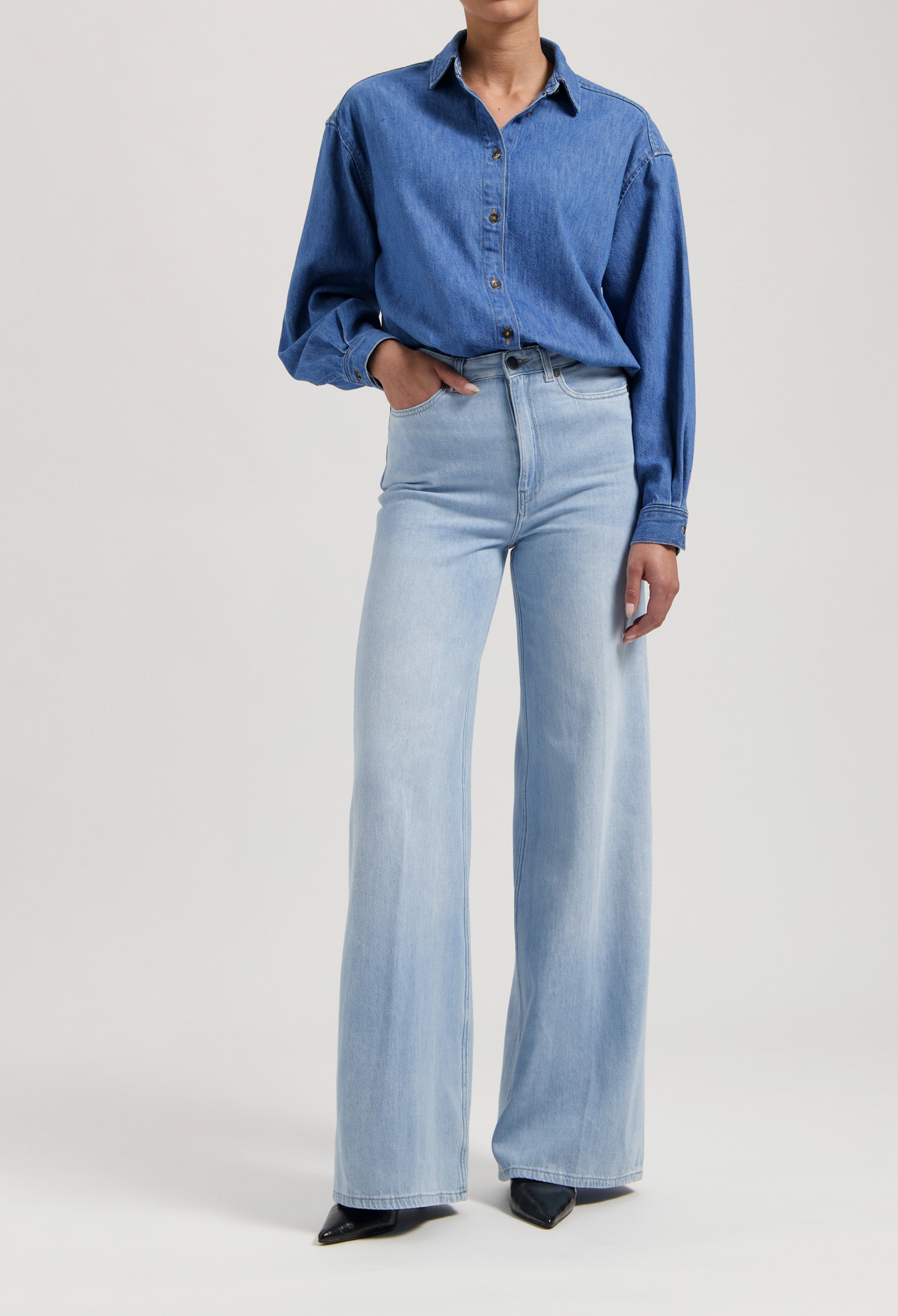 Woman wearing a blue denim shirt and high-waisted light blue wide-leg jeans with hands in pockets, featuring black pointed shoes.