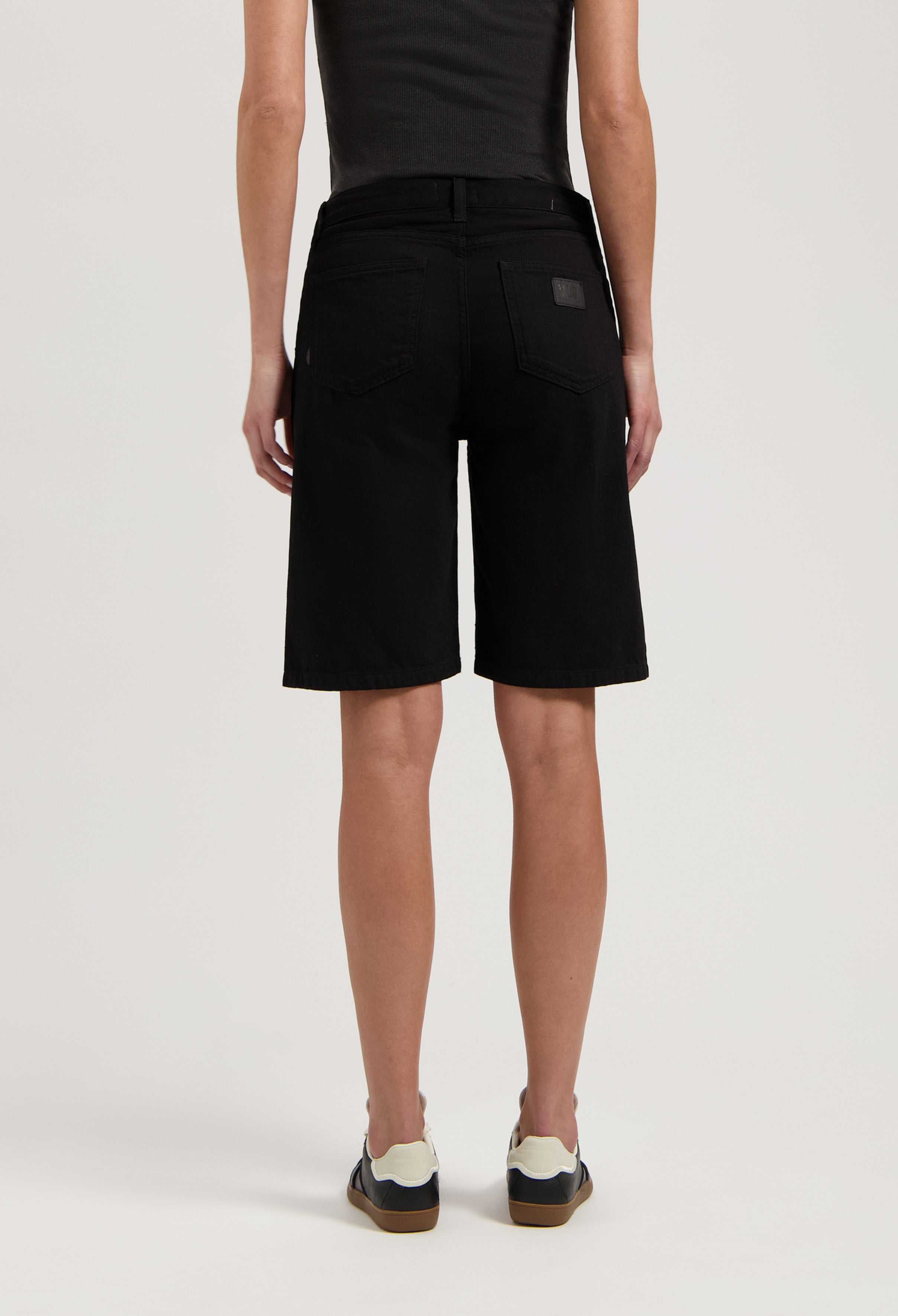 Black denim Bermuda shorts made from cotton, showing a back view worn with a black top and white sneakers.