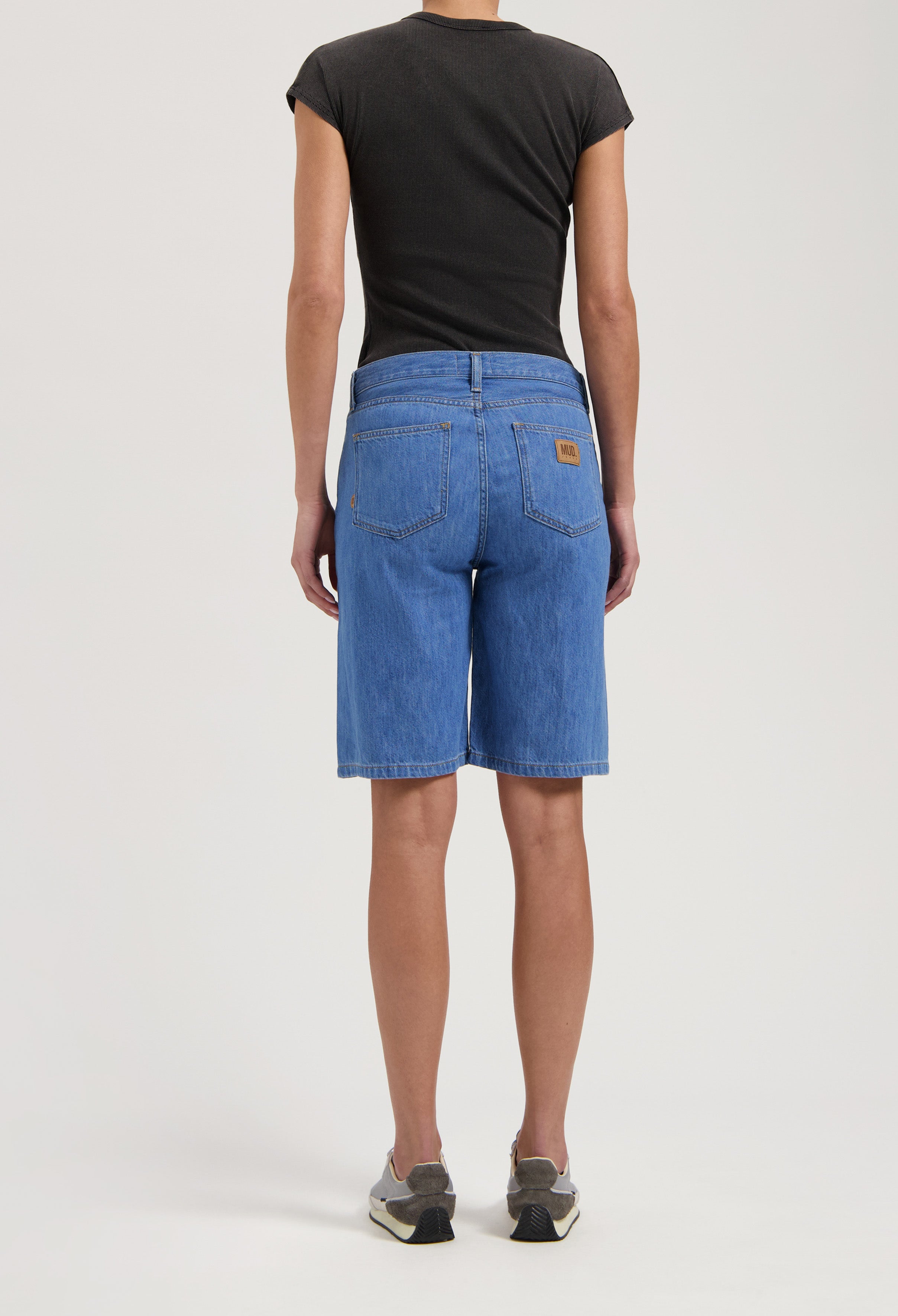 Back view of blue knee-length denim shorts by MUD Jeans, showing a casual style paired with a fitted black t-shirt.