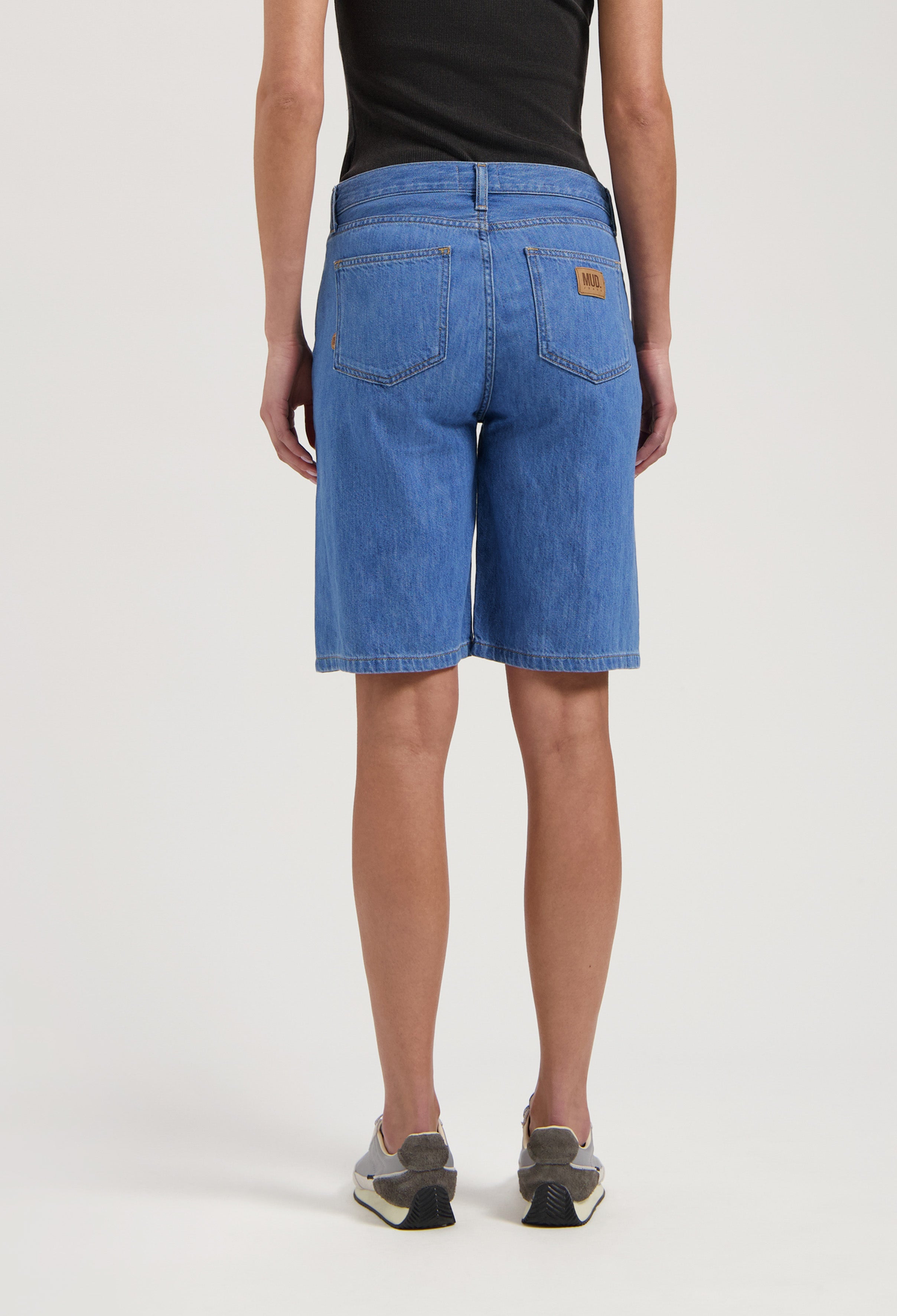 Mid-length eco-friendly MUD Jeans in blue denim, featuring a relaxed fit, worn with a black tank top and athletic shoes.