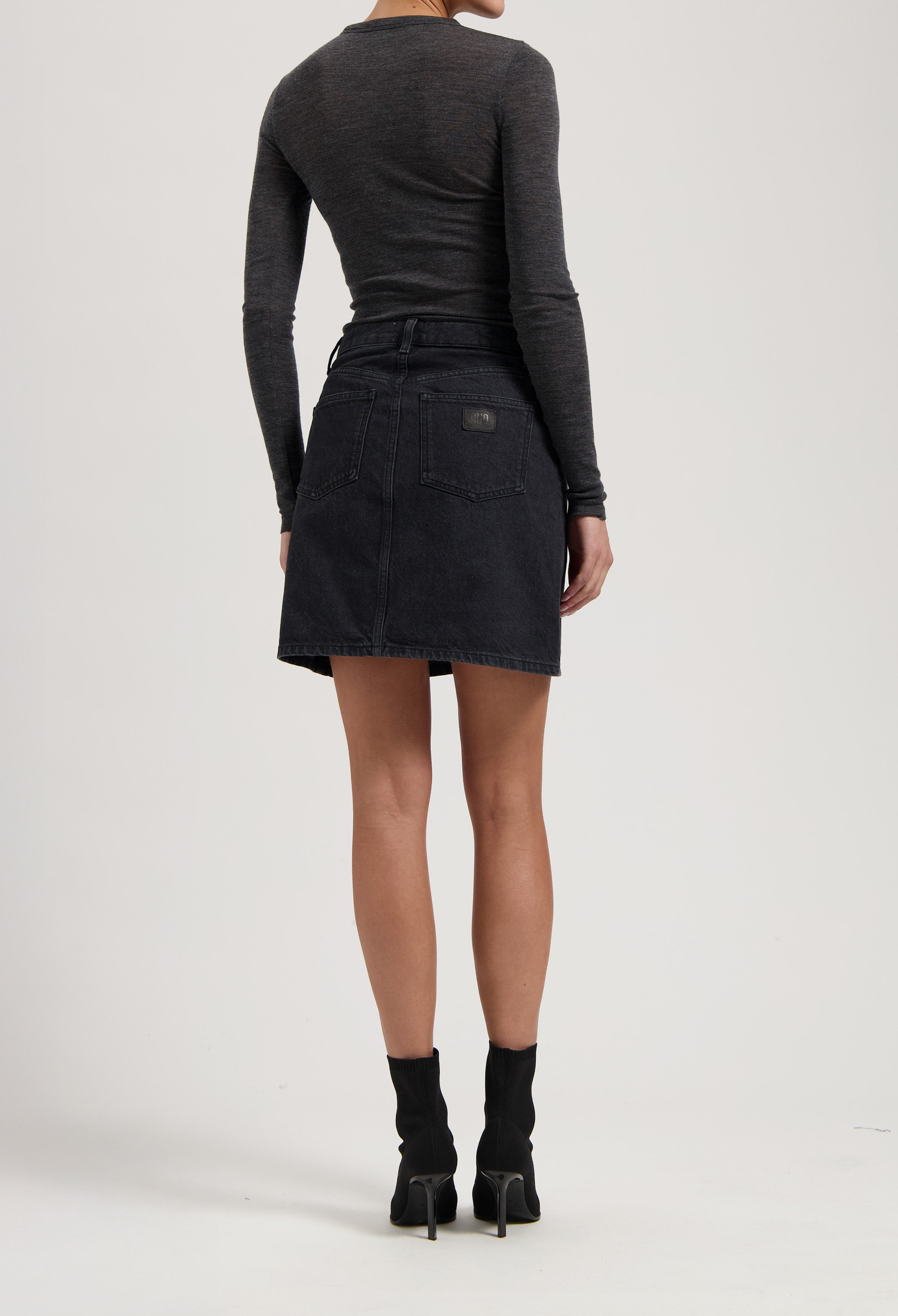 Back view of a model in a dark gray long-sleeve top and black denim skirt, paired with black heeled ankle boots.