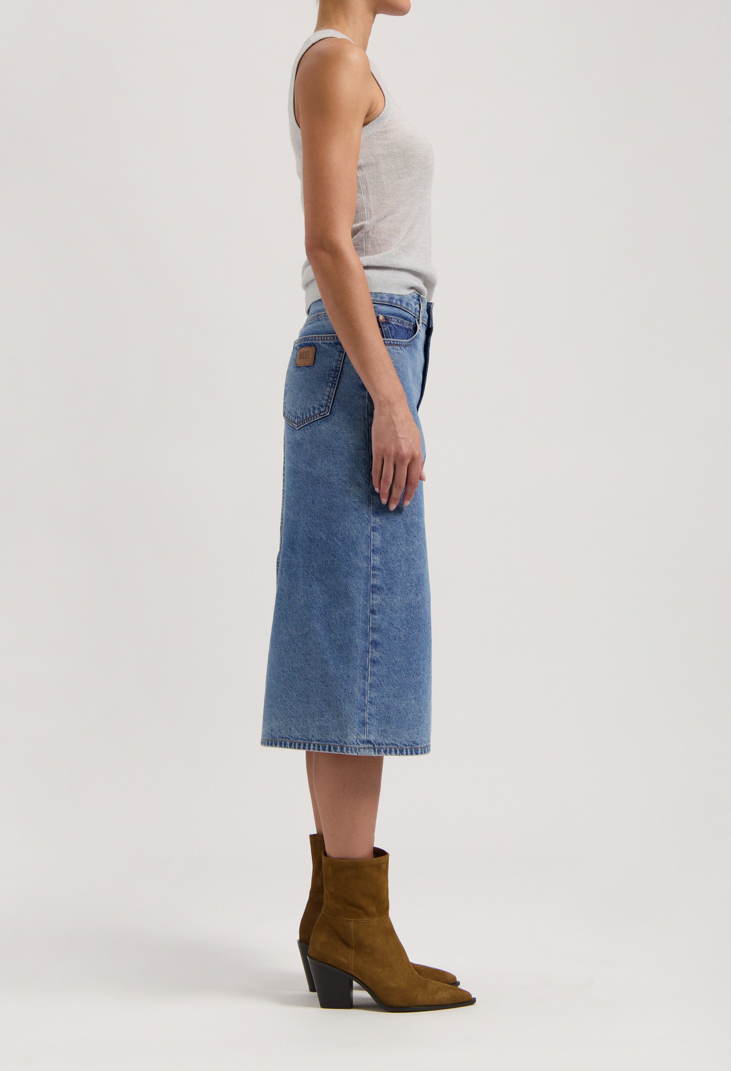 Side view of a woman in a sleeveless gray top, wearing a blue denim midi skirt paired with brown suede ankle boots.