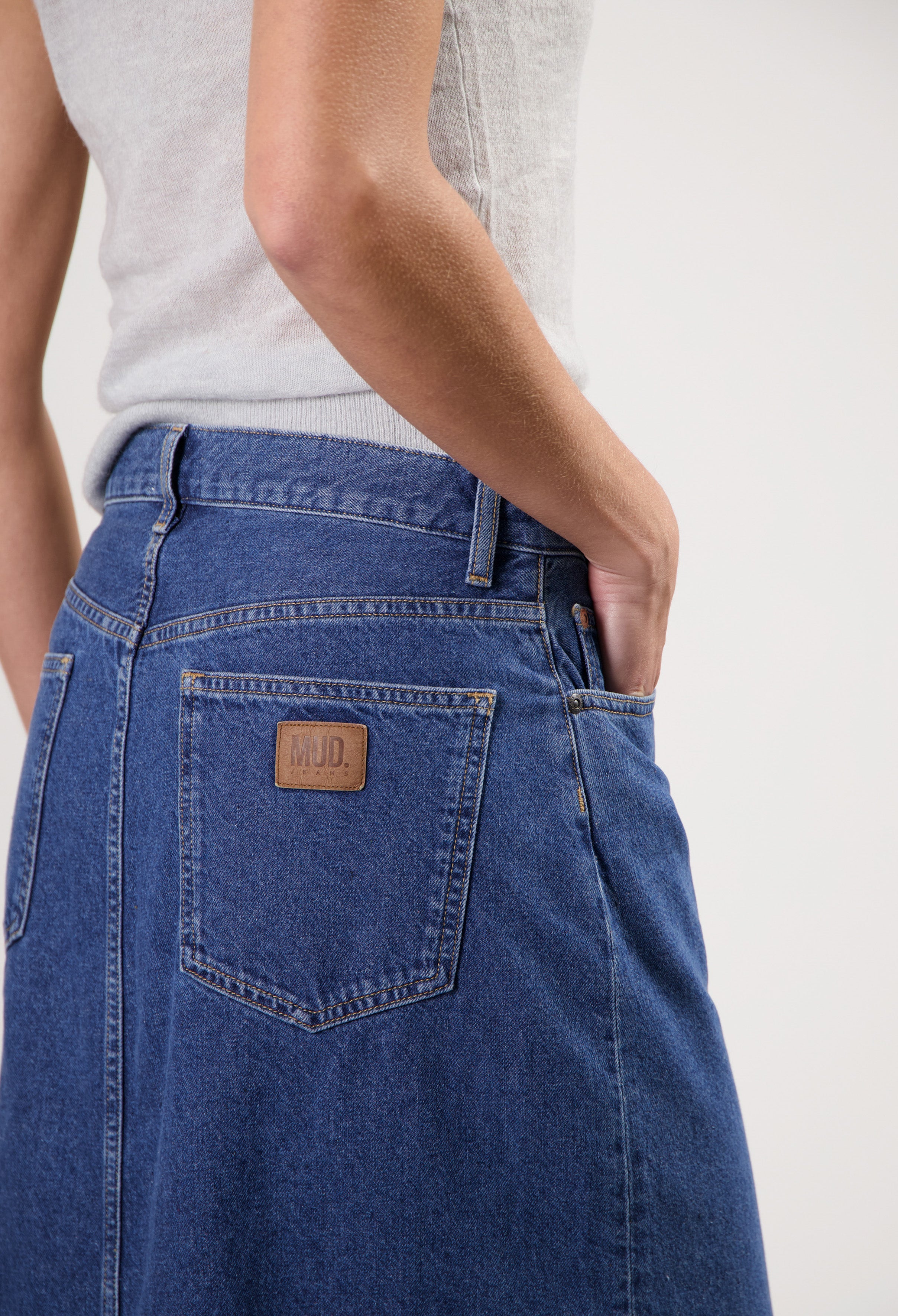 Close-up of a model wearing blue MUD Jeans denim skirt with visible pocket stitching and leather patch, paired with a white tank top.
