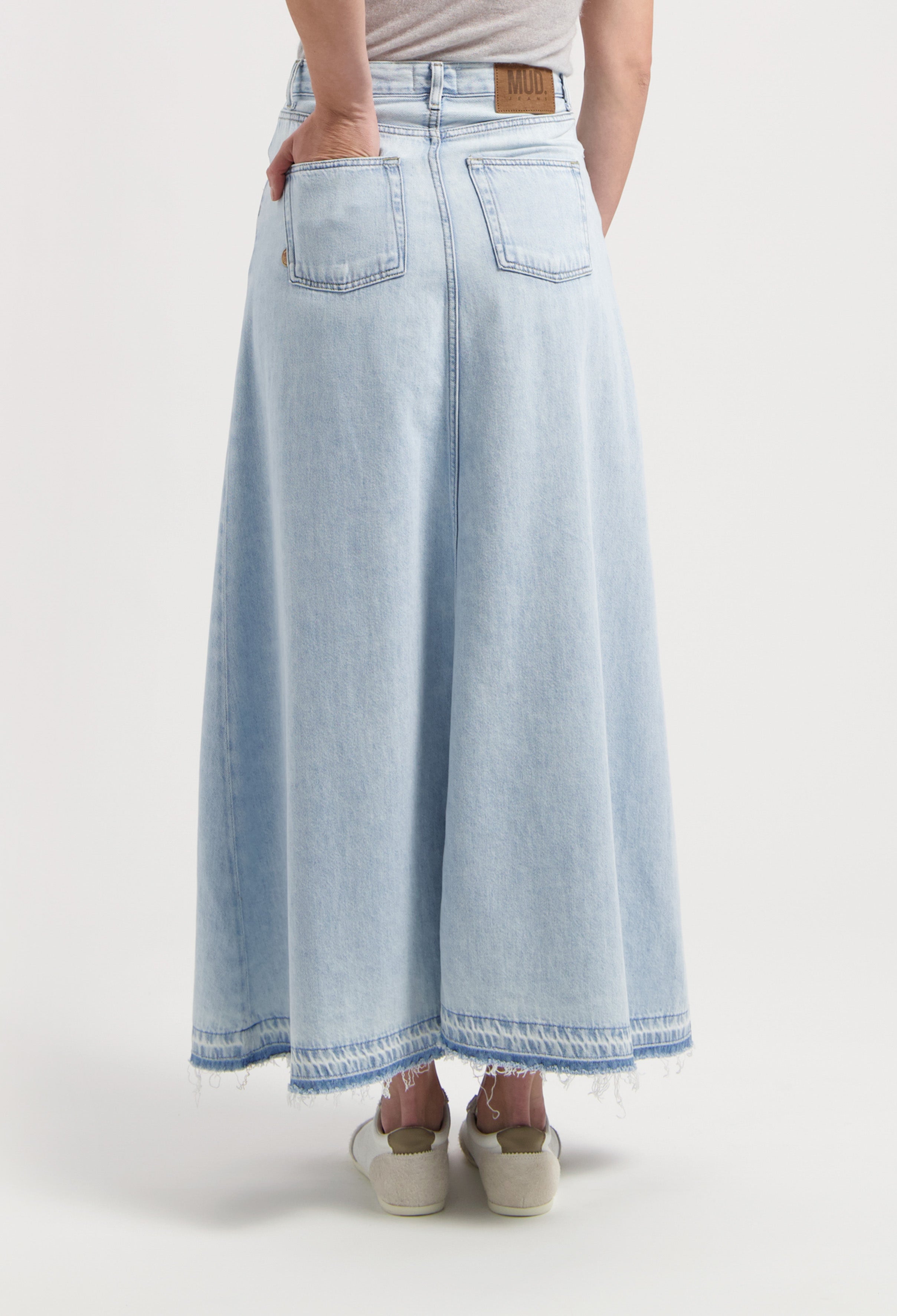 Light blue, ankle-length denim skirt by MUD Jeans, showcasing back pockets and a frayed hem, paired with casual shoes.