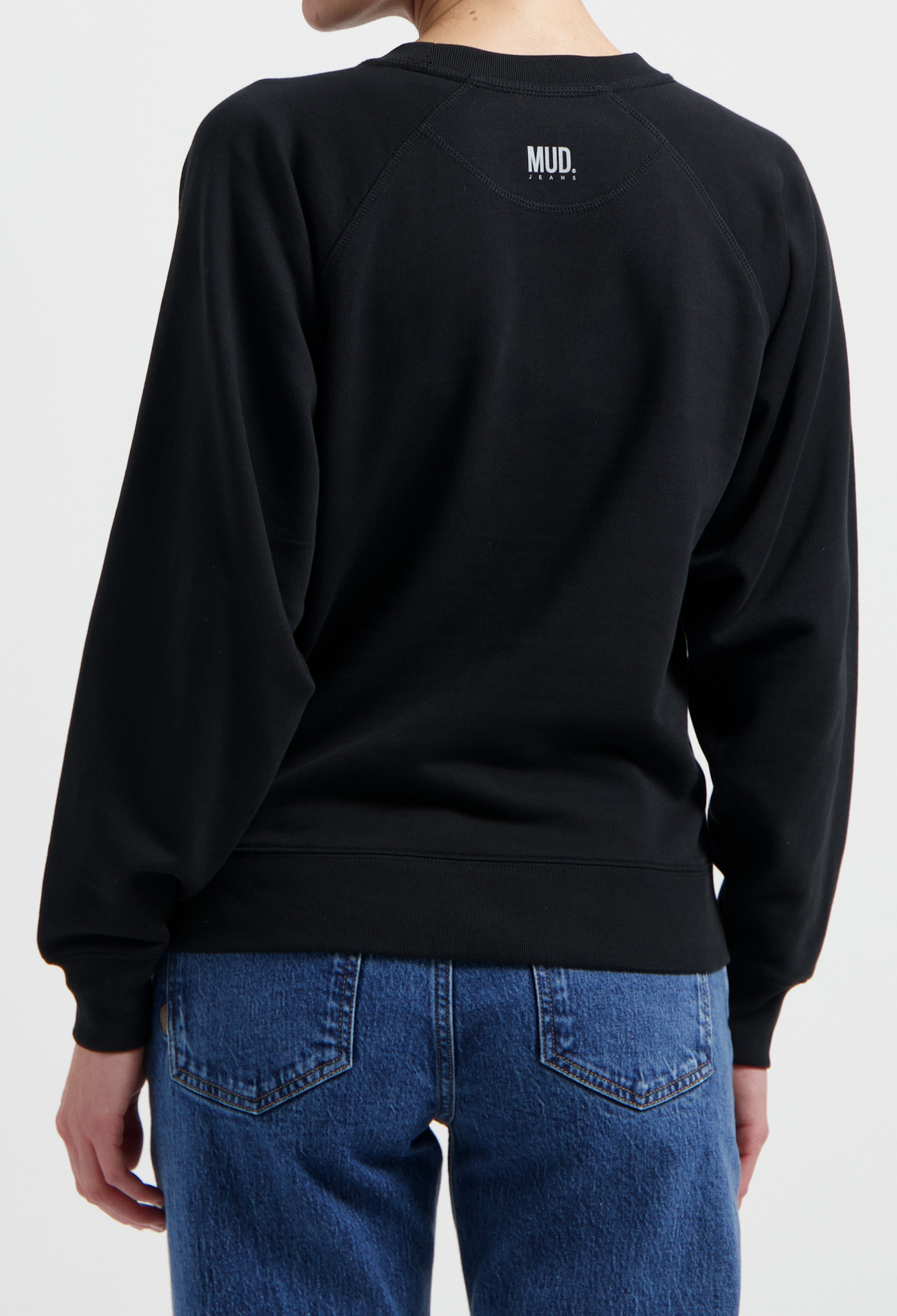 Black MUD Jeans sweatshirt with simple logo, paired with blue denim jeans, showcasing casual and comfortable style.
