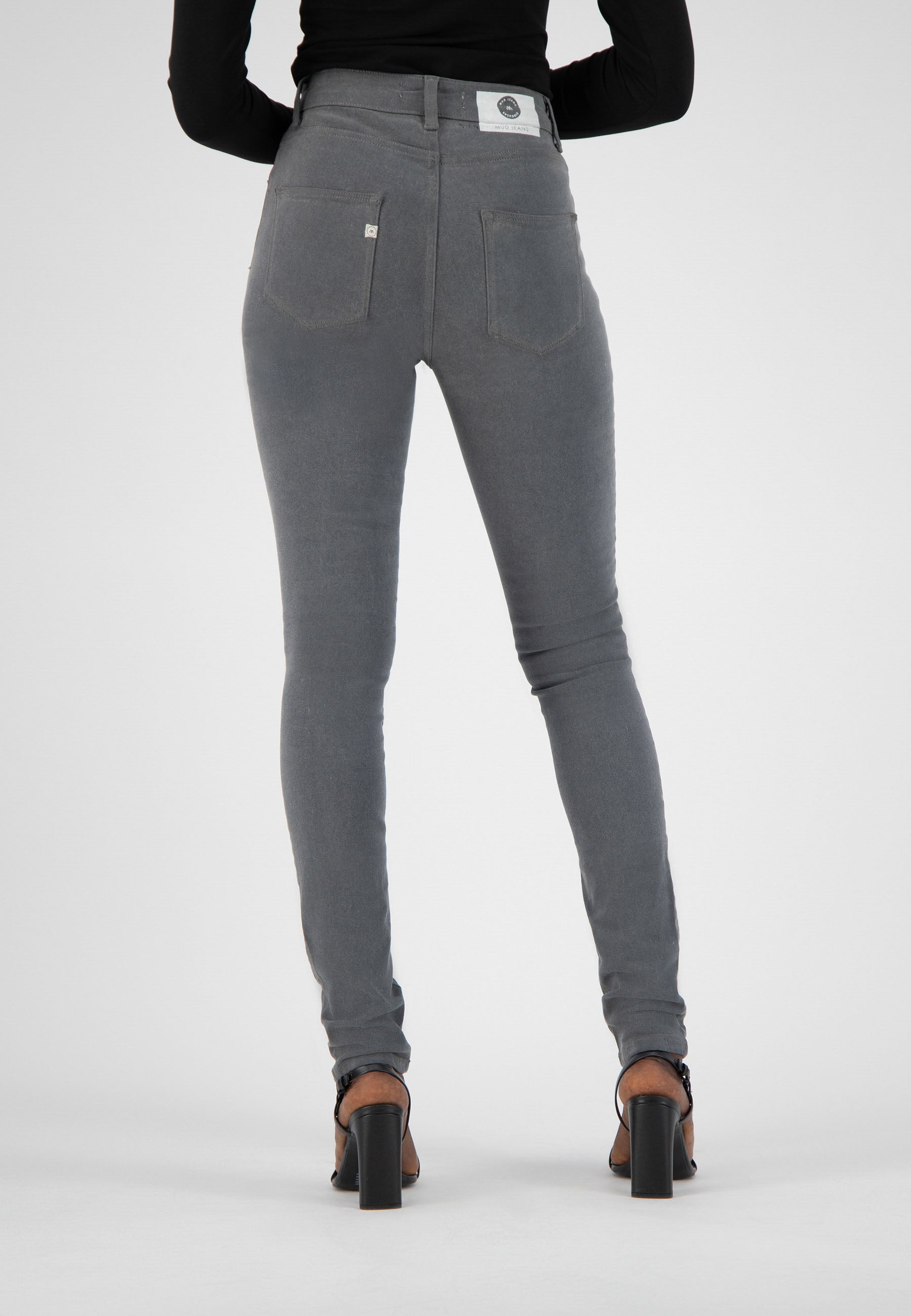 Gray slim-fit denim jeans from MUD Jeans, high-waisted design, paired with a black long-sleeve top and black heels.