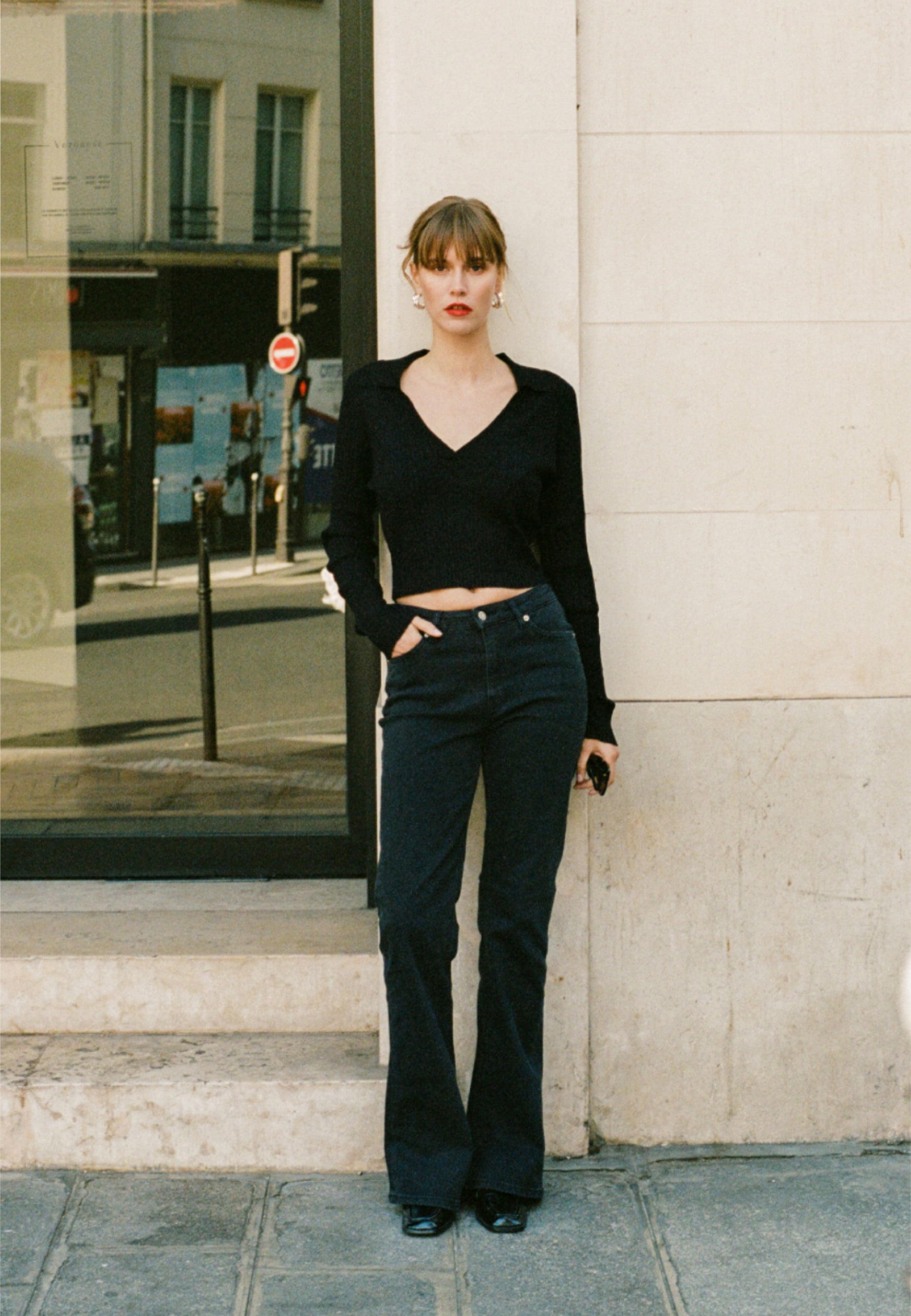Woman in a black V-neck sweater and dark flared jeans, standing confidently against a light stone wall in urban setting.